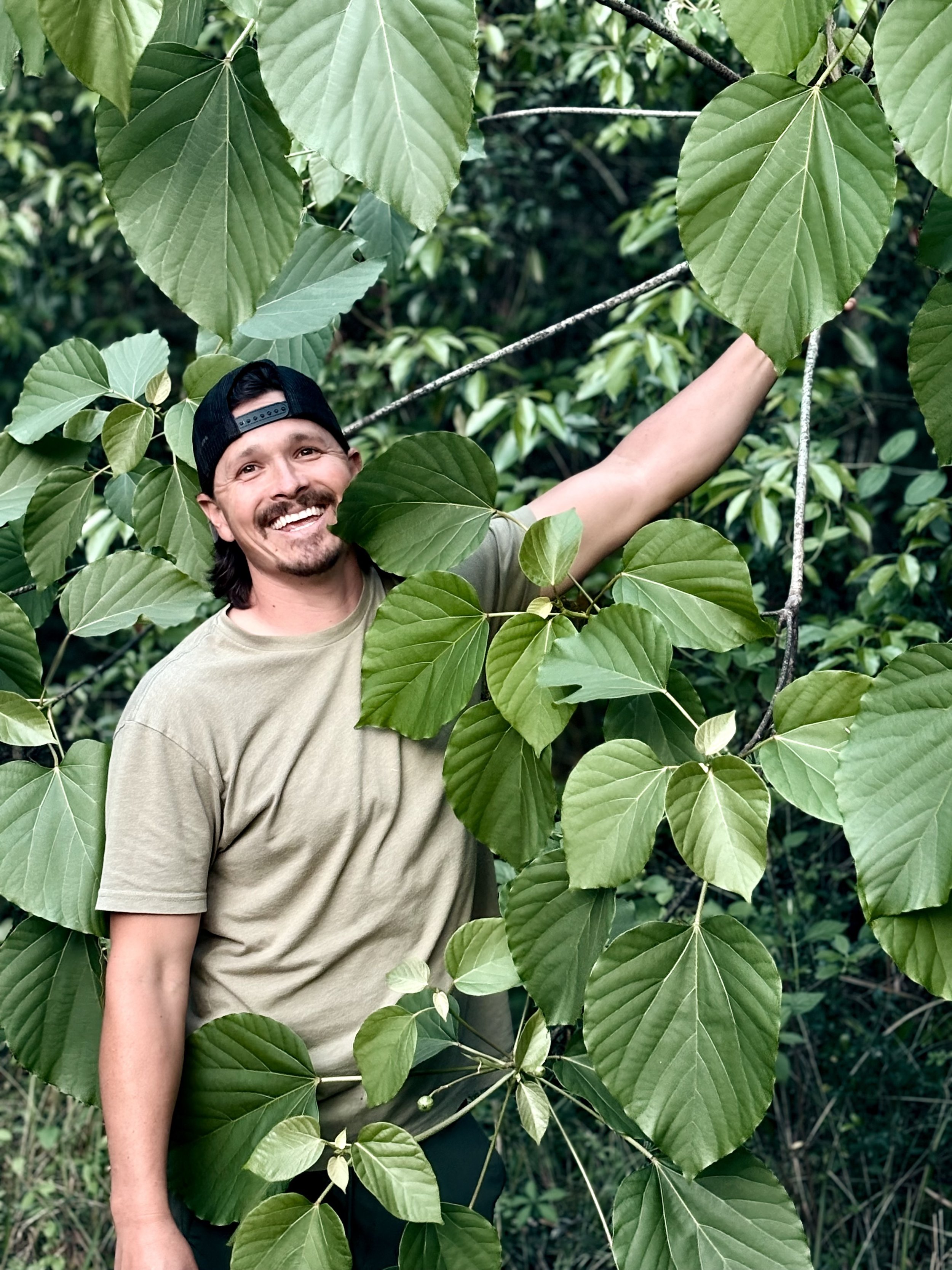 A man smiling and wearing a beige t-shirt and a black cap, standing among lush green leaves in a forest or jungle.