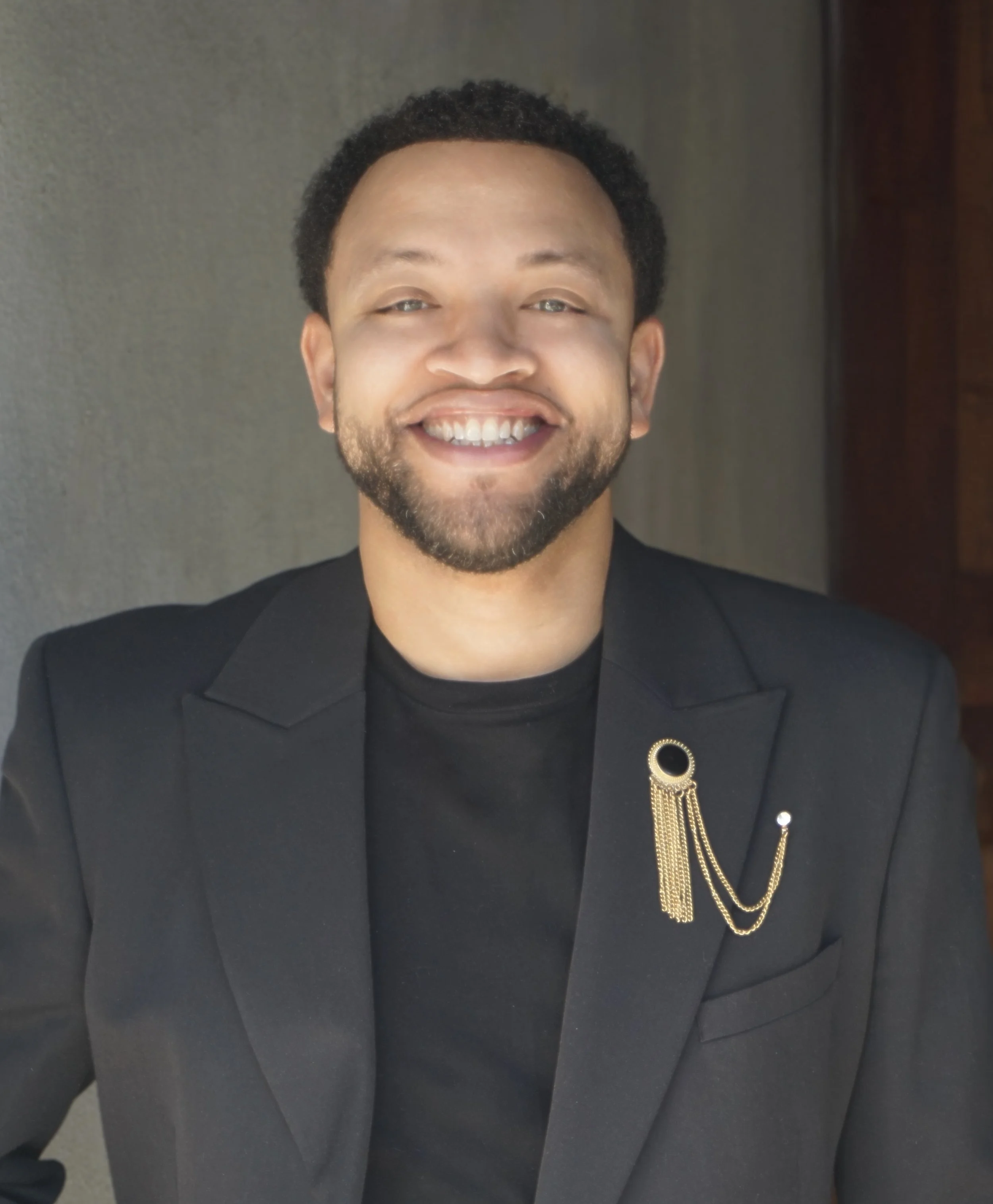 Close-up of a man smiling, wearing a black tuxedo with a decorative gold chain on the lapel.
