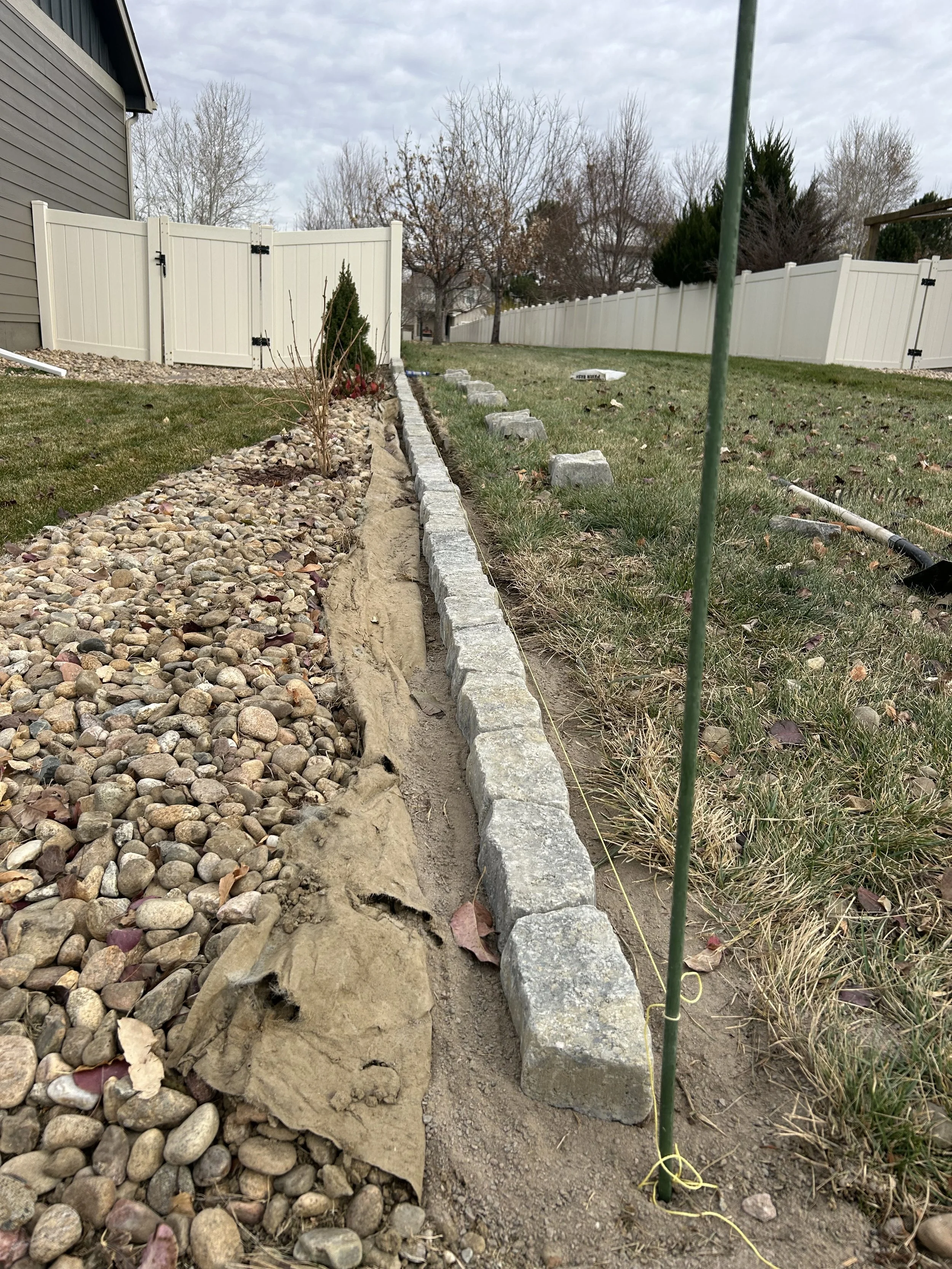 A backyard with a stone and gravel border, some plants, and a white fence in the background.