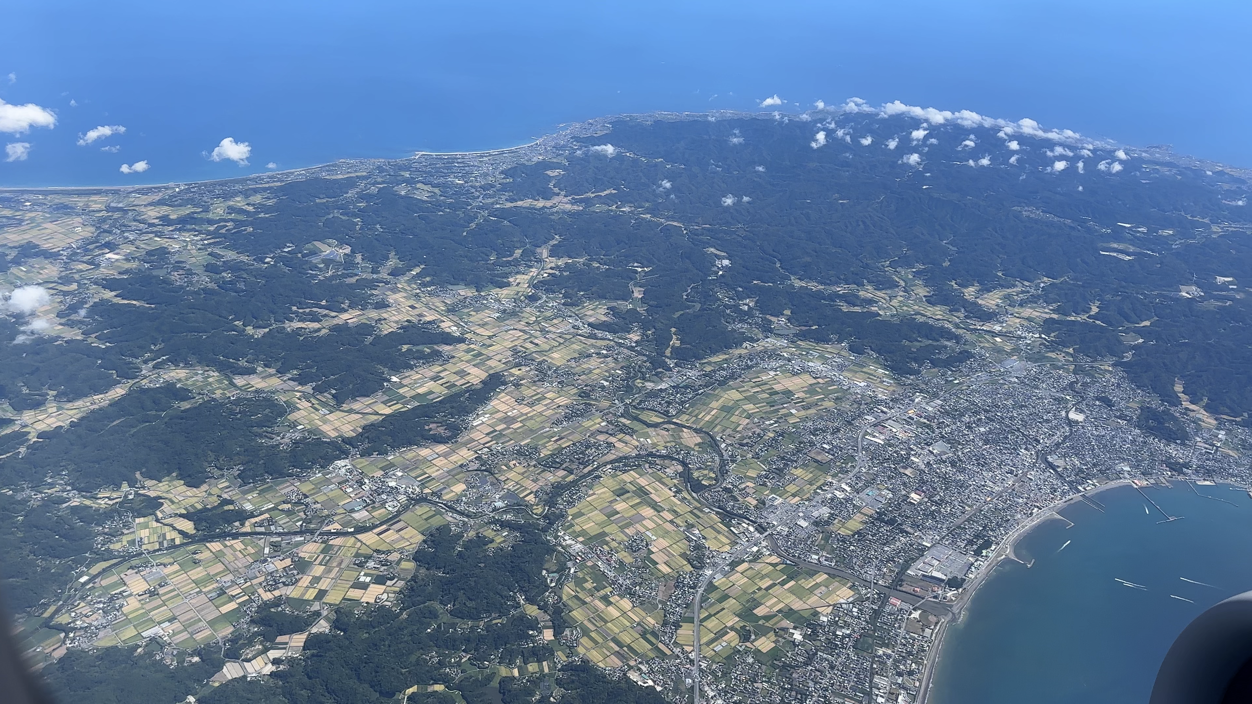 Aerial view of a coastal city with a mix of urban and rural areas, fields, hills, and the ocean in the background.