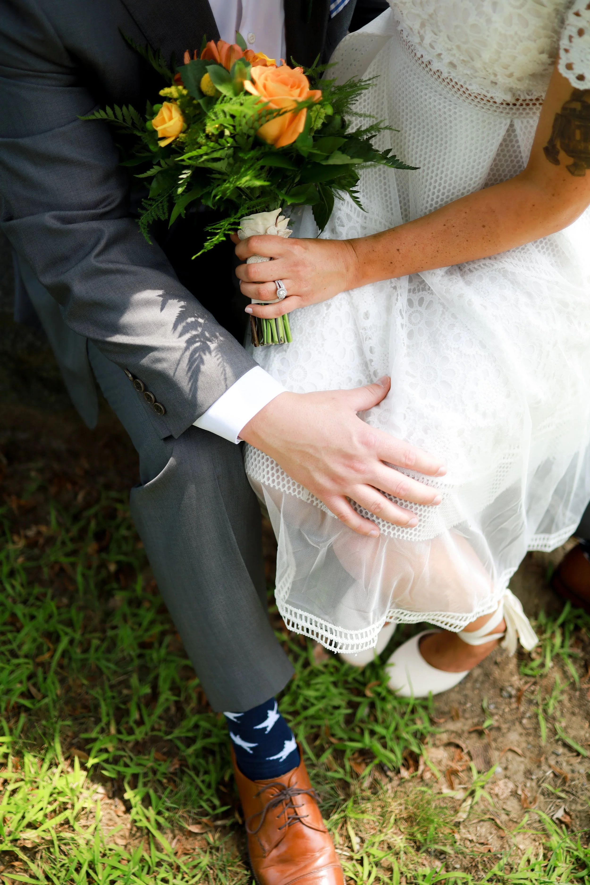 A bride and groom holding a bouquet of orange and yellow flowers together. The groom wears a gray suit with dark socks featuring white swans, brown shoes, and a white shirt. The bride is dressed in a white lace dress, showing a ring on her finger and has a tattoo on her arm. They are standing on grass and dirt outdoors.