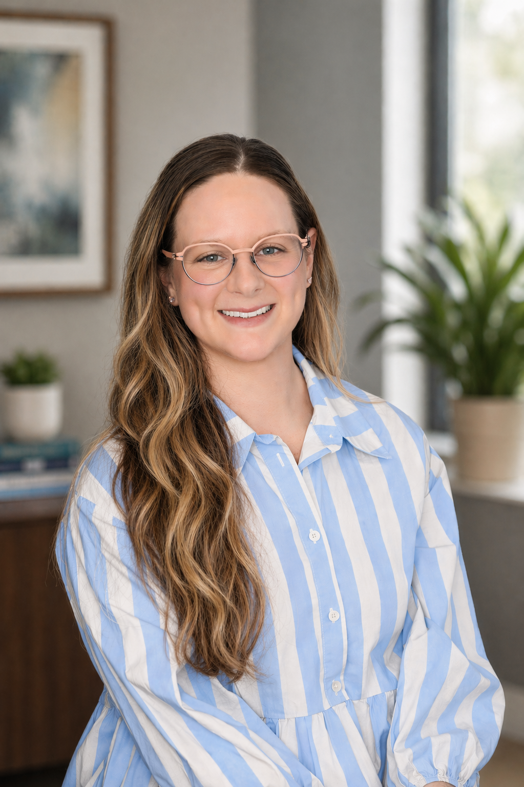 A woman with long wavy brown hair, wearing glasses and a blue-and-white striped shirt, smiling in a well-lit room with plants and artwork in the background.