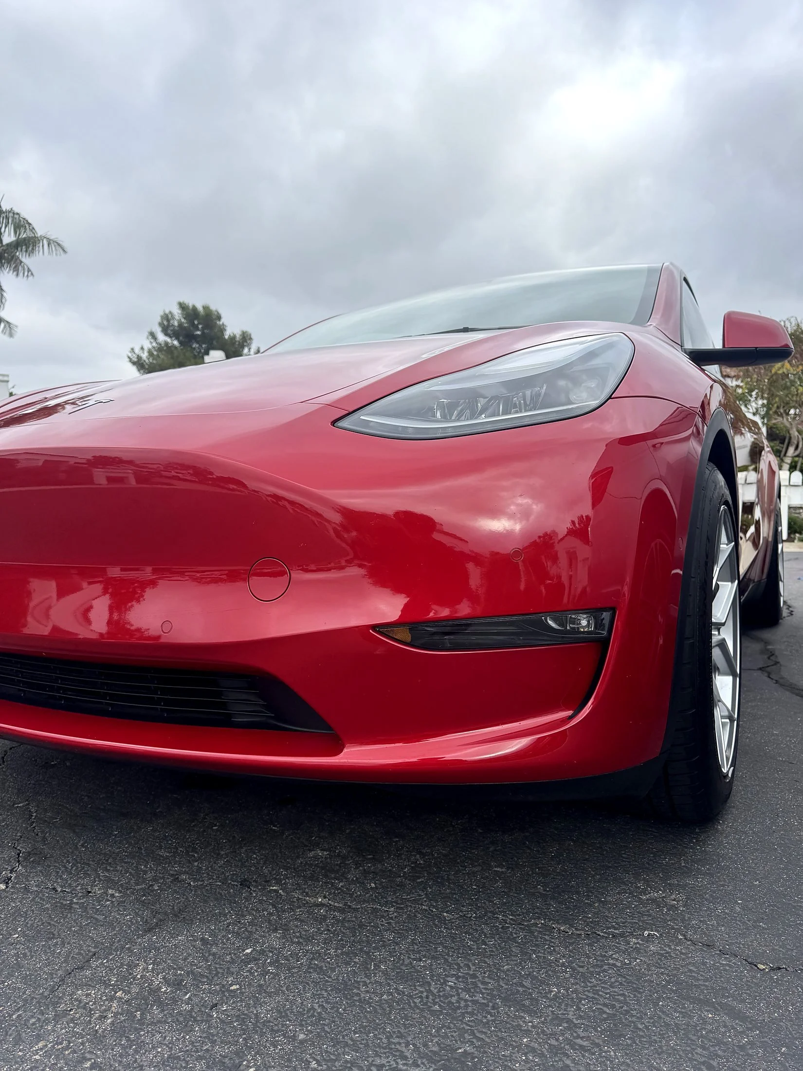Close-up of a red sports car, showing the front front-left side, with sleek curves, headlight, and wheel, parked on street with cloudy sky overhead.