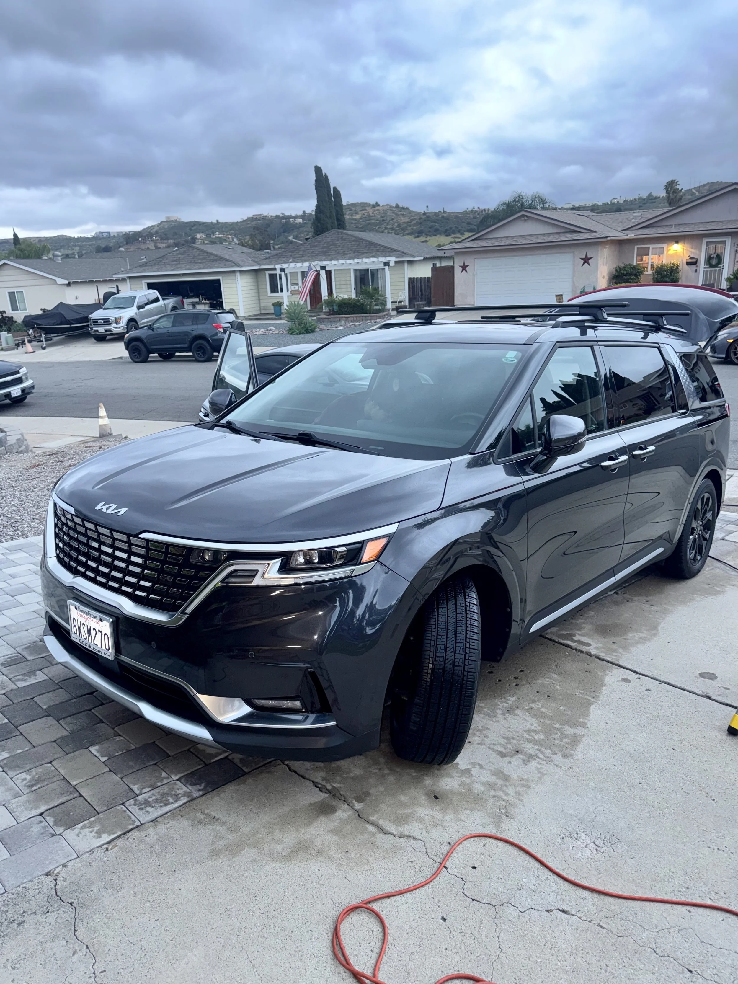 A residential driveway with multiple parked cars, including a black Kia SUV in the foreground, a silver truck and a gray SUV, and a boat on a trailer. The sky is overcast, and there are hills with houses and trees in the background. An orange extension cord is on the driveway, which is cracked and uneven.