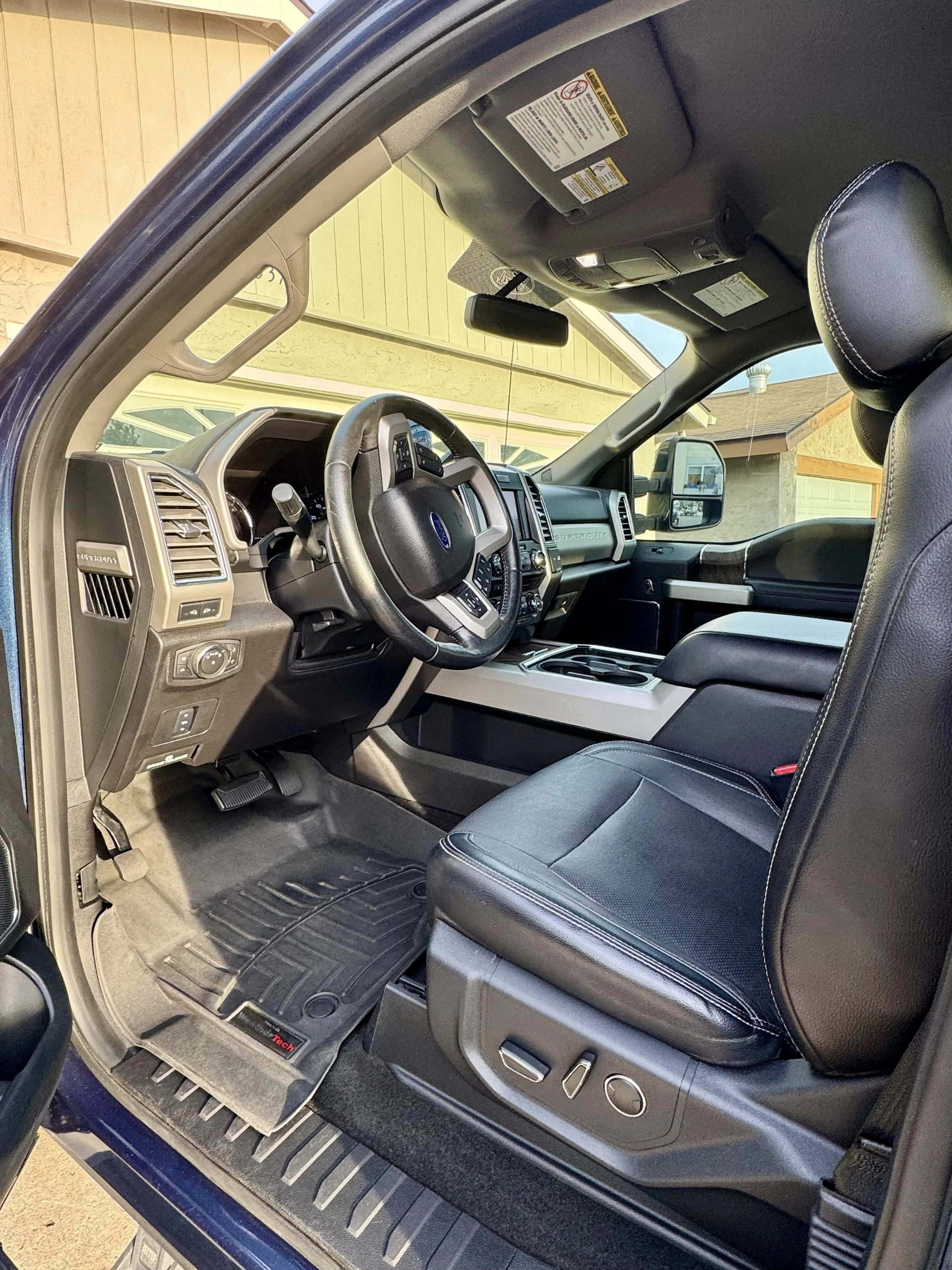 The interior of a pickup truck, showing the driver's seat, steering wheel, dashboard, and part of the front passenger seat. The floor mat and door panel are also visible. The exterior of a house is seen through the window.