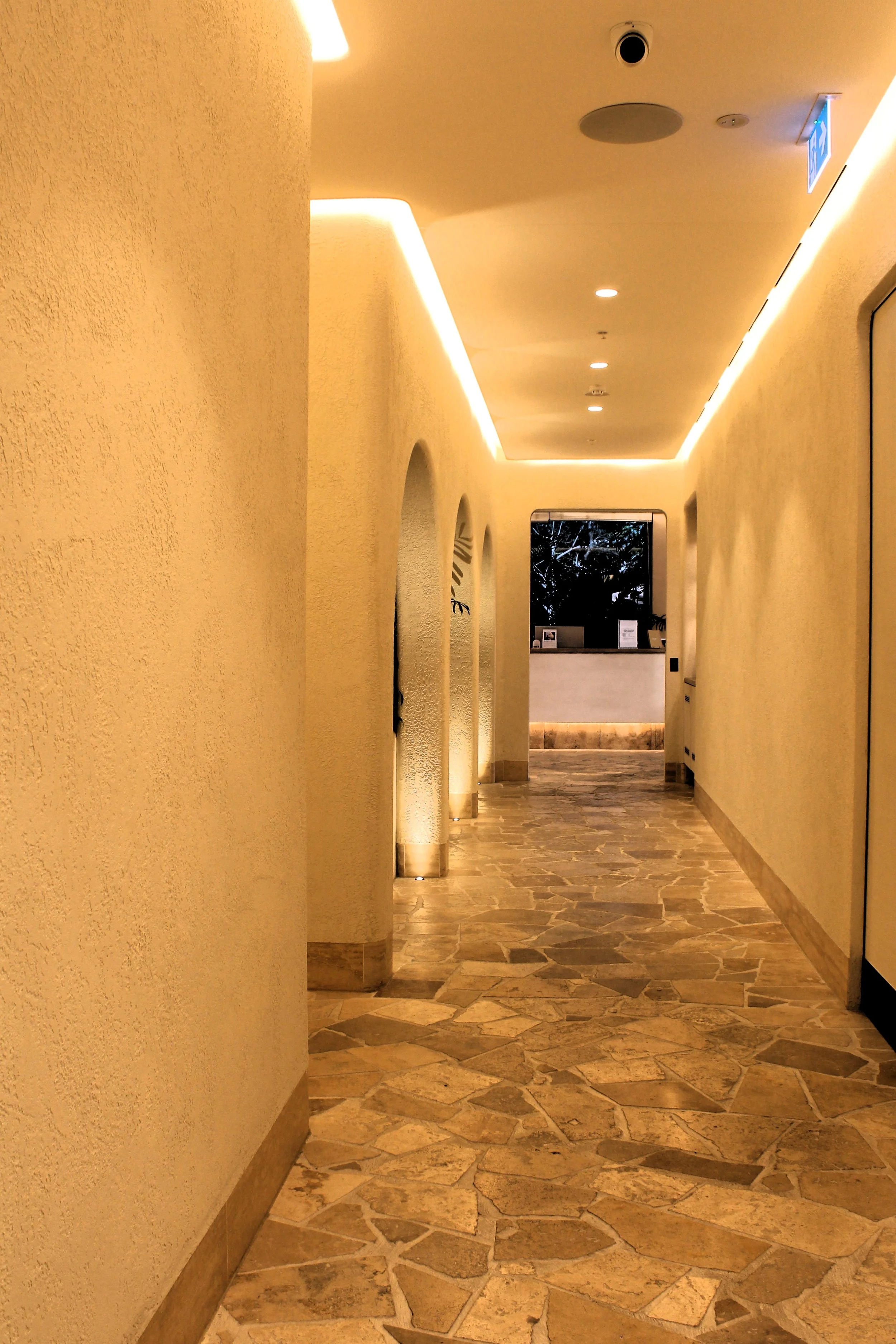 Long hallway with beige walls, archways, and wooden flooring tiles, illuminated by warm lighting and a window at the end showing trees outside.