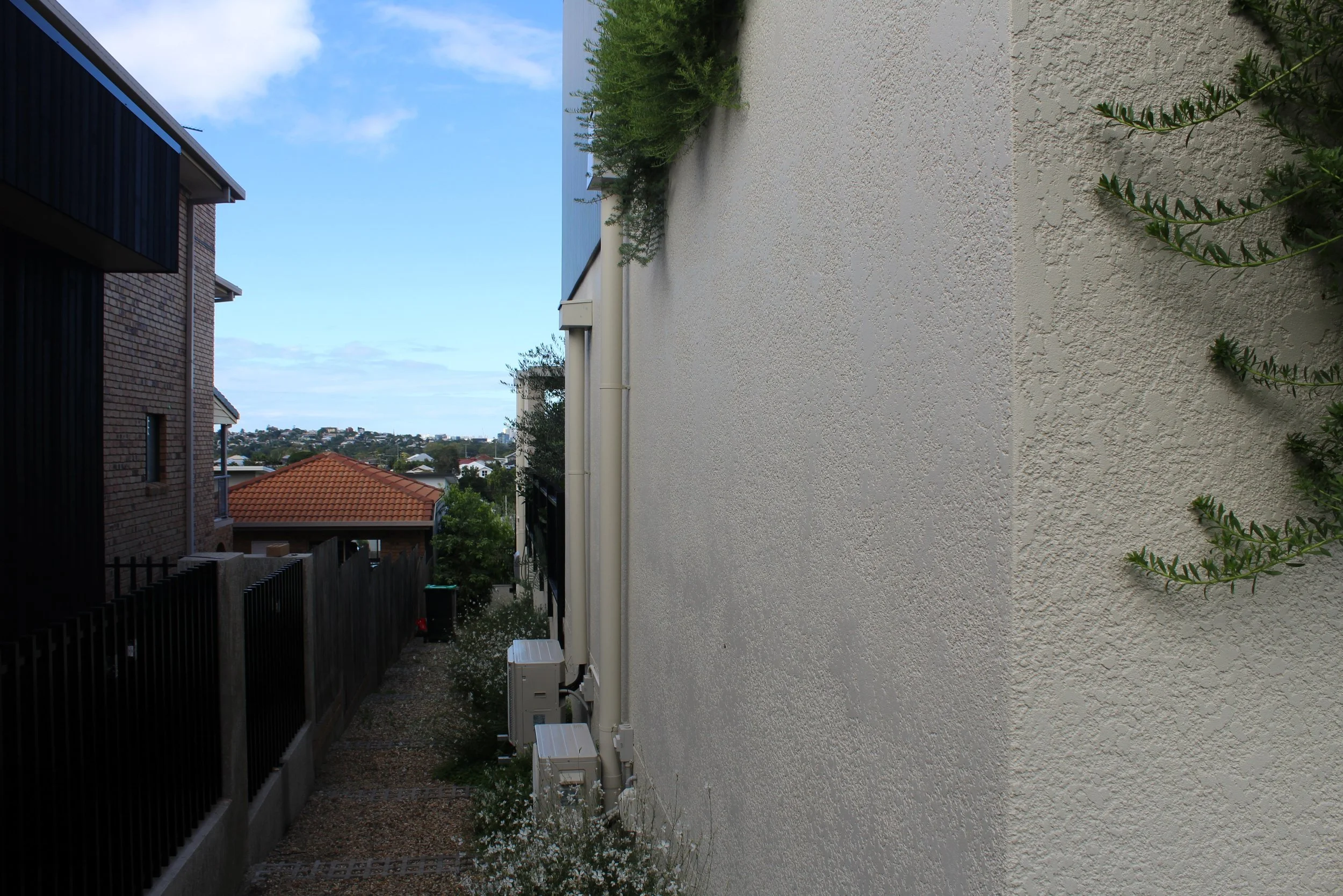 Side view of a house exterior showing a white textured wall with air conditioning units and pipes, neighboring house with brick and black siding, a fence, and a view of rooftops and trees under a blue sky with clouds.