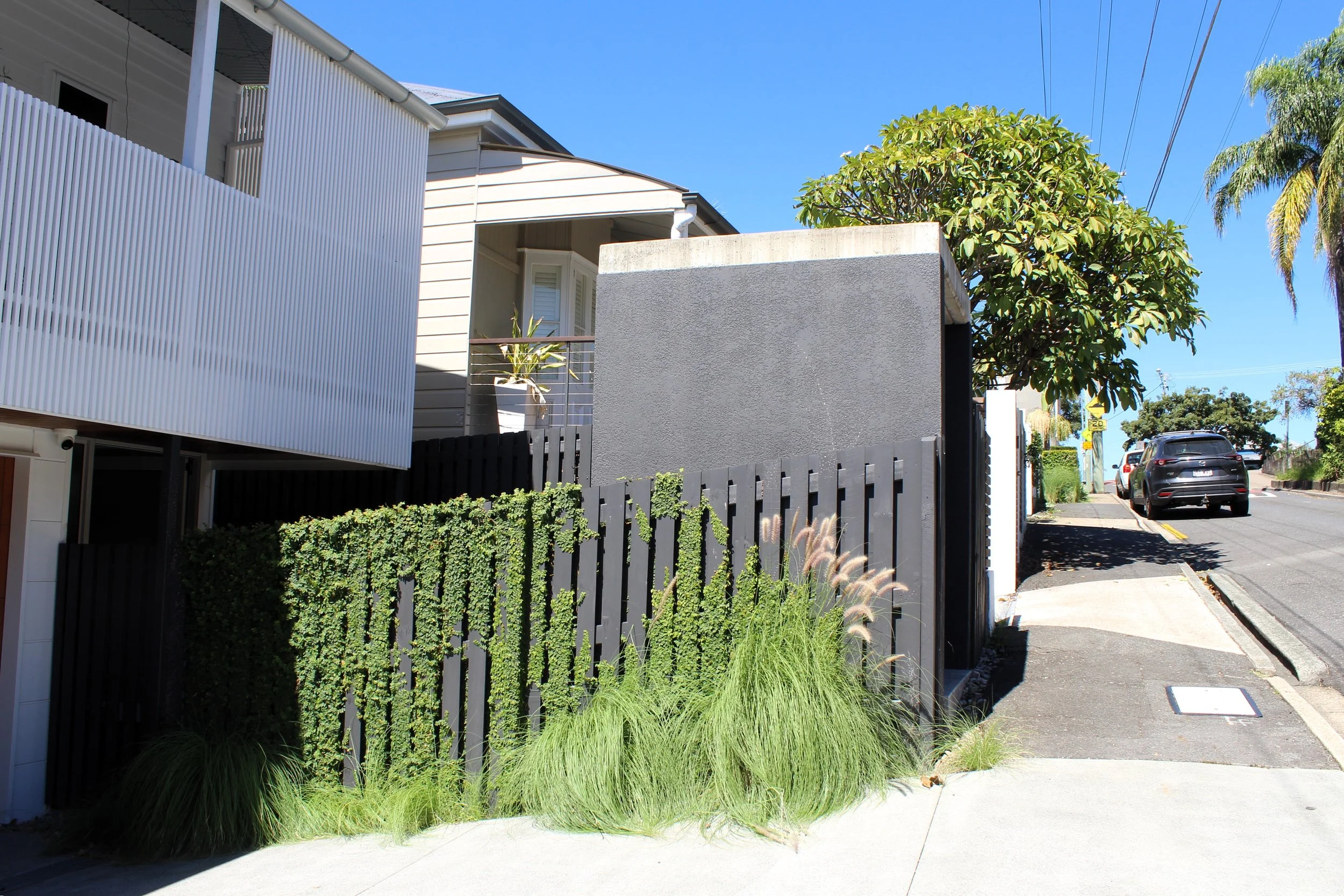 Residential street scene with modern house, gray wall, black fence, green plants, blue sky, parked cars, and trees in the background.