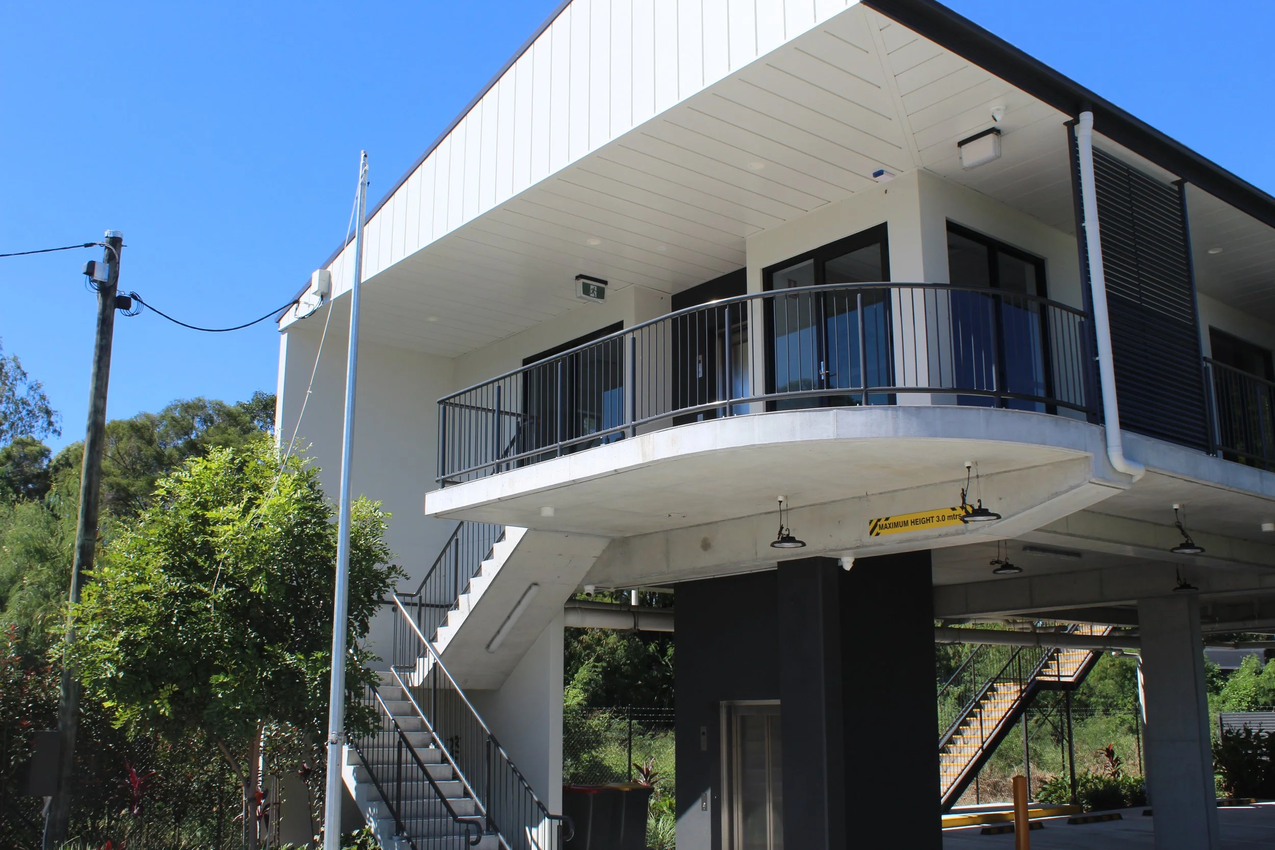 Modern multi-story building with balcony, stairs, and parking area beneath, surrounded by greenery and trees.