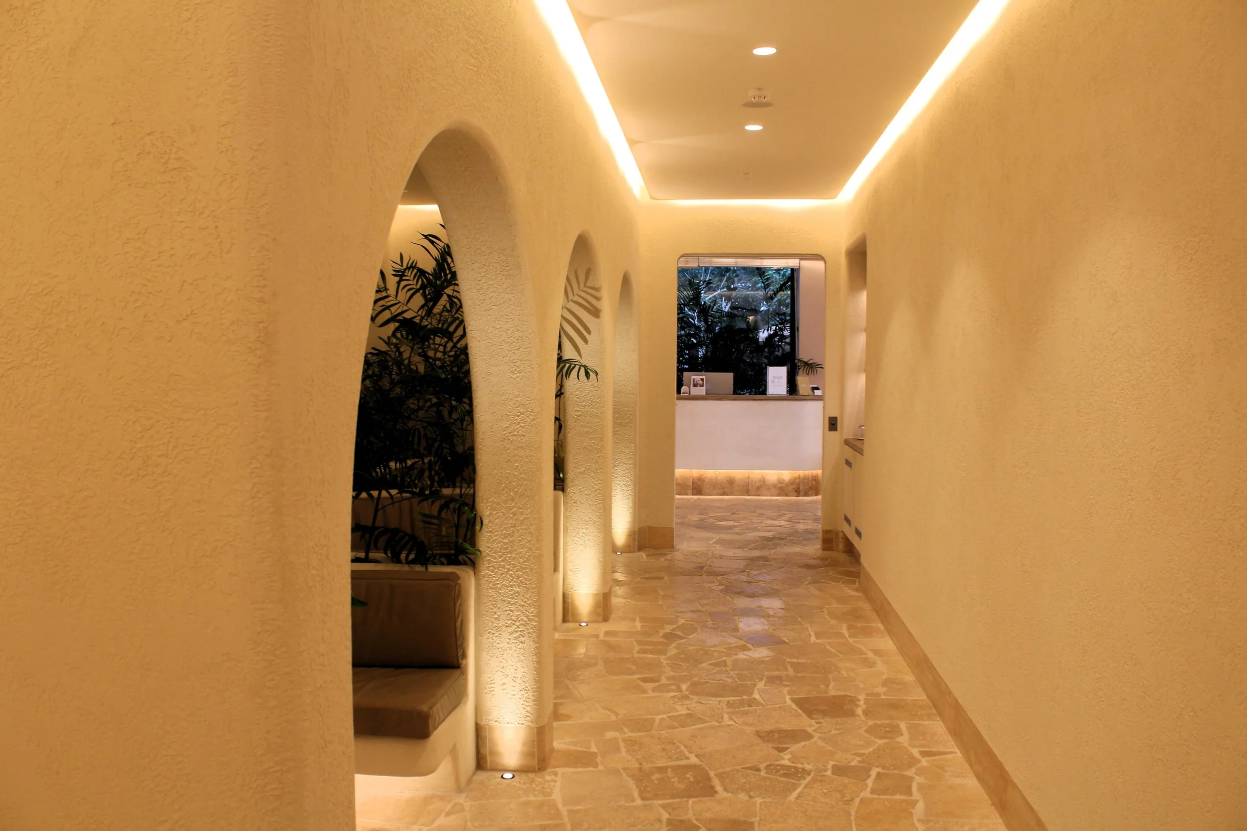 A warm-colored hallway with textured yellow walls, arched wall cutouts with built-in benches, and a tile floor. The ceiling has recessed lighting and crown molding with cove lighting. At the end of the hallway, there is a reception desk with a large 