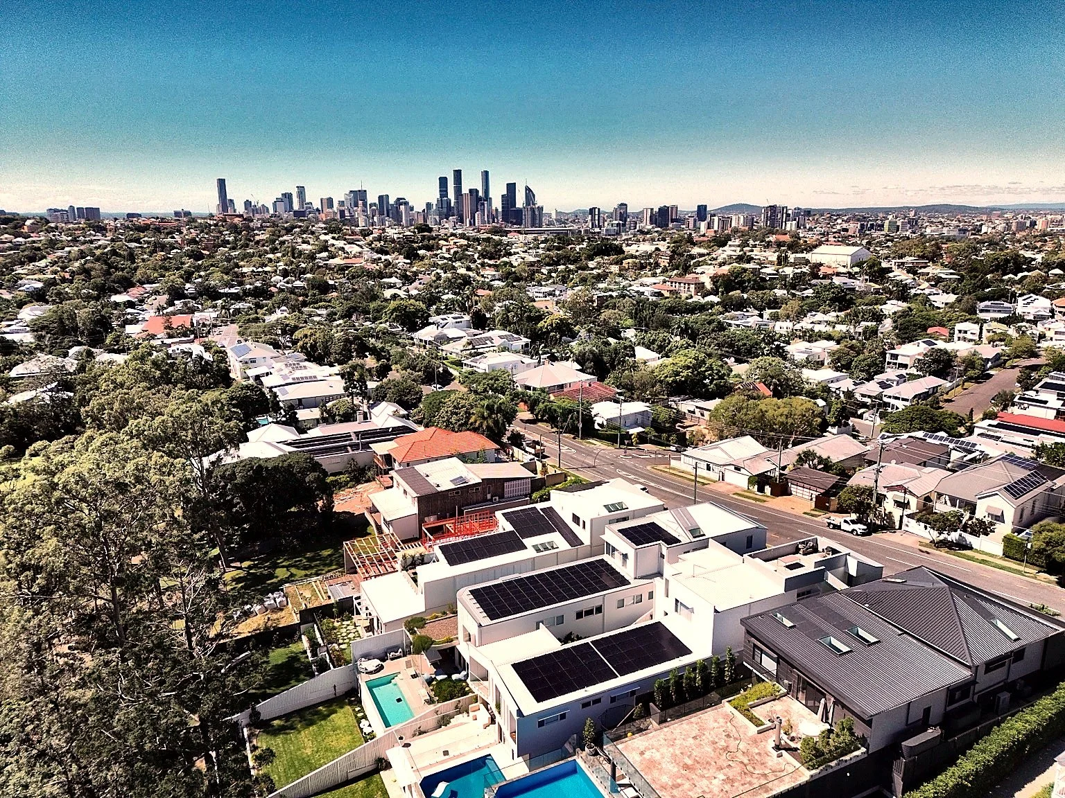 Aerial view of a suburban neighborhood with modern houses, swimming pools, and solar panels, with a city skyline in the distance under a clear blue sky.