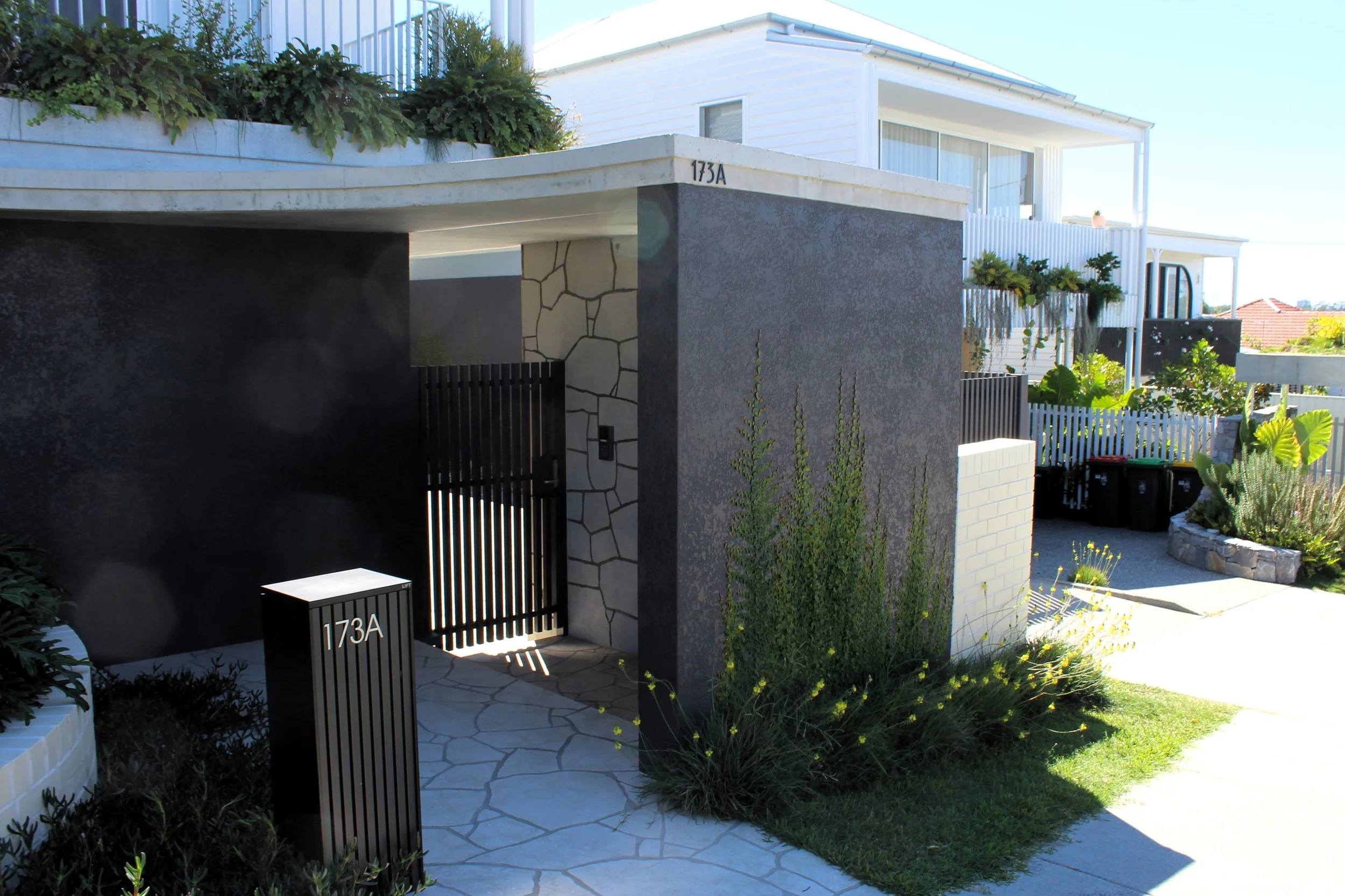 Front entrance to a modern house with white exterior walls and large windows, featuring a black gate with the number 173A, surrounded by lush greenery and patio plants, with a stone walkway leading to the door and a sunny sky overhead.