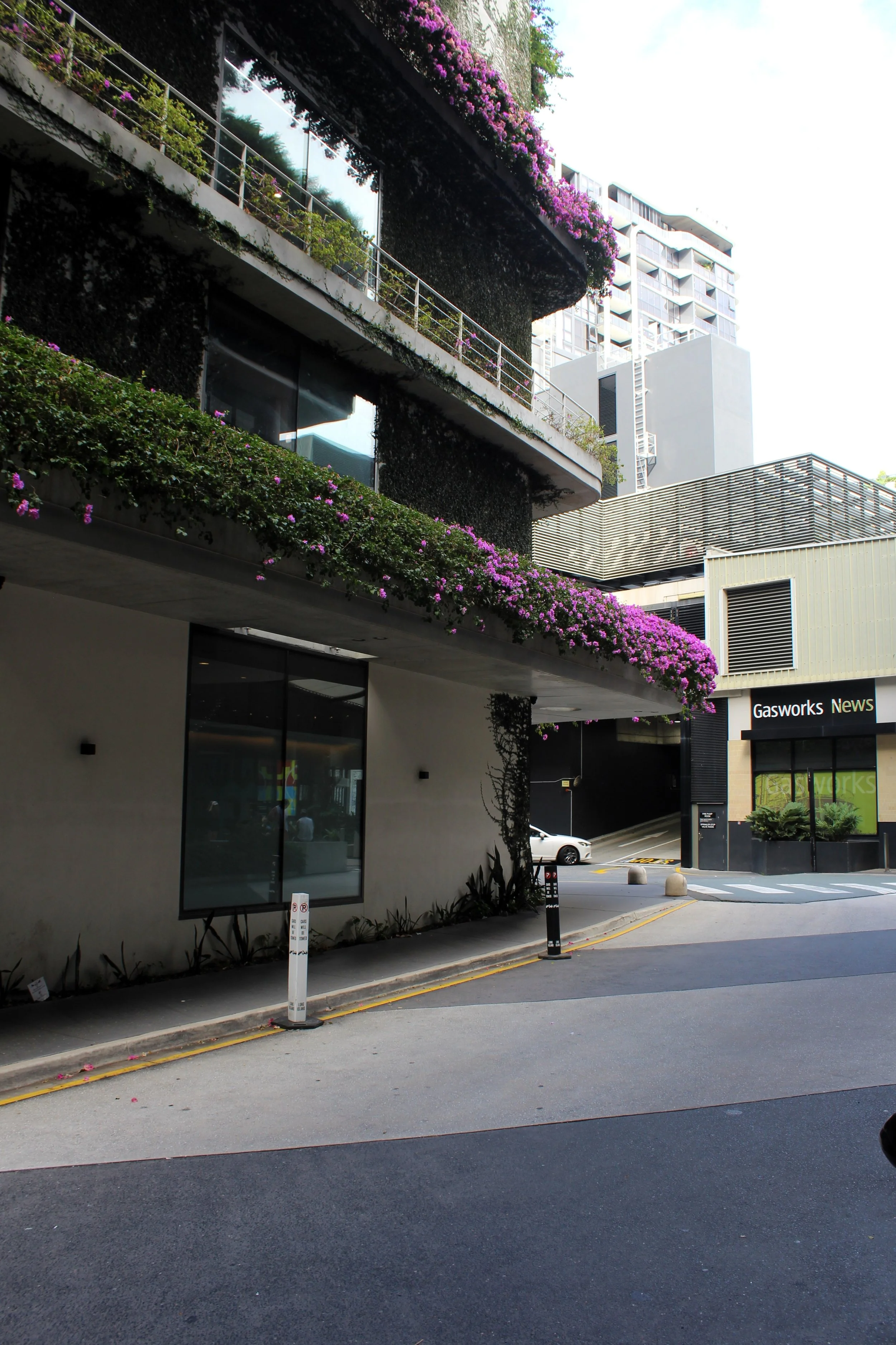 A modern building with lush green plants and pink flowers on the balconies, located in an urban area with a parking lot and a street in front.