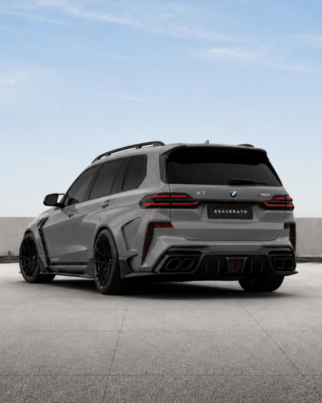 Rear view of a matte gray Esagerato BMW X7 M60i SUV with black wheels, sporty body kit, and quad exhaust, parked on a concrete surface with a clear sky background.