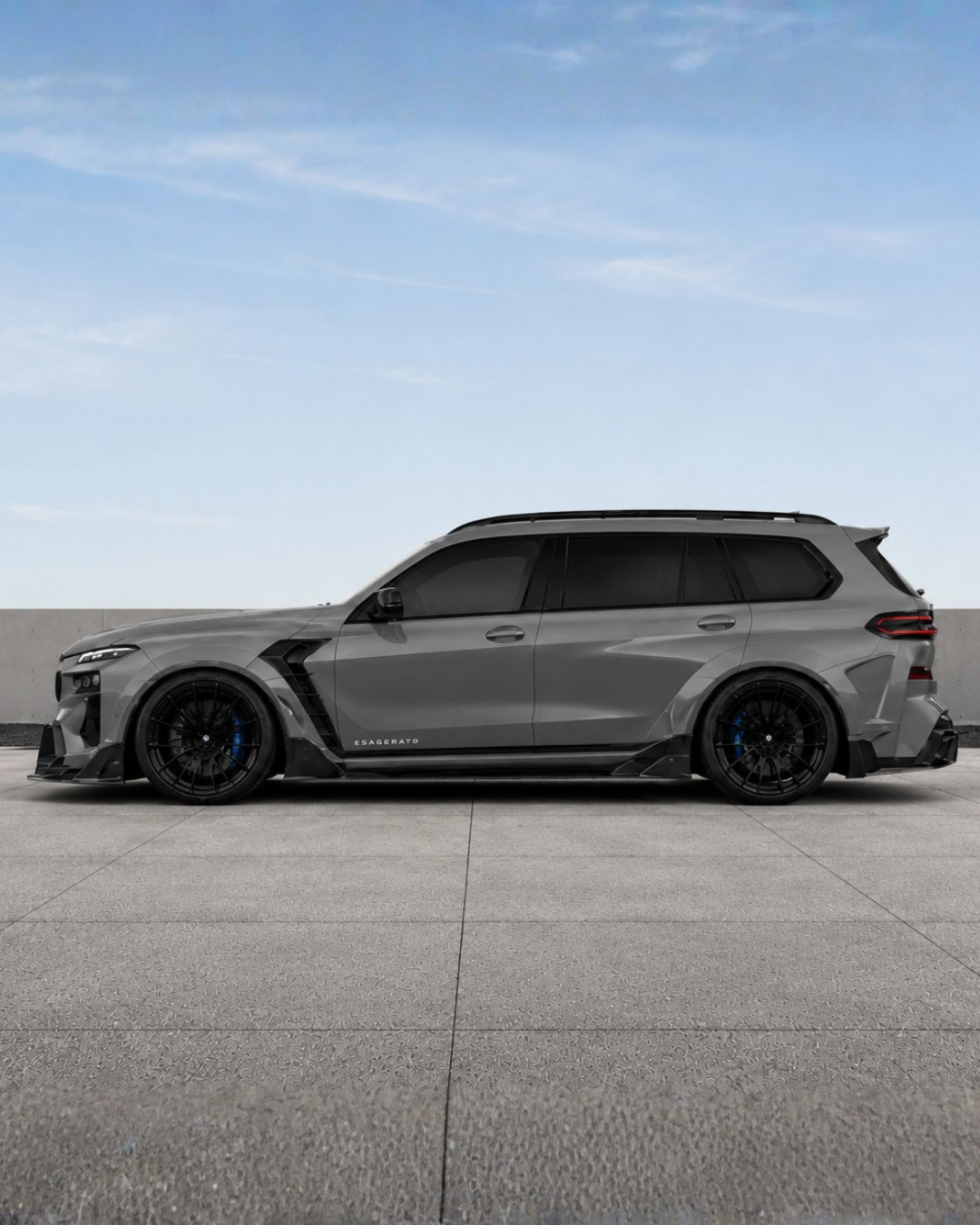An Esagerato BMW grey luxury SUV with custom black wheels and body kit parked on a rooftop with a concrete wall and blue sky in the background.