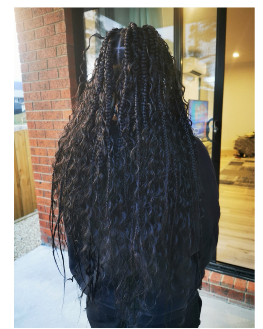 Person with long curly braided hairstyle standing outside near a brick wall and glass door.