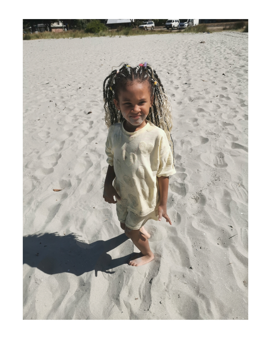 A young girl with braided hair and colorful beads, wearing a light yellow shirt and shorts, standing barefoot on sandy ground on a sunny day at the beach.