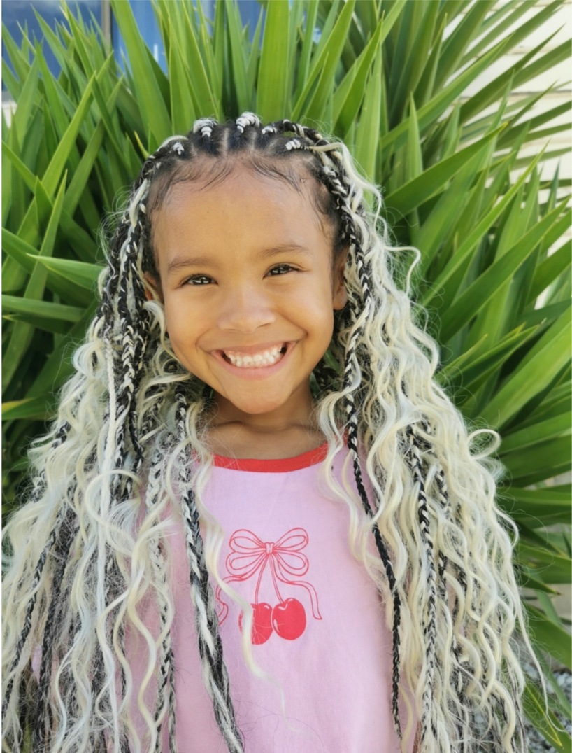 A smiling girl with long, curly, blonde hair with black and white braids standing outdoors in front of large green leaves.