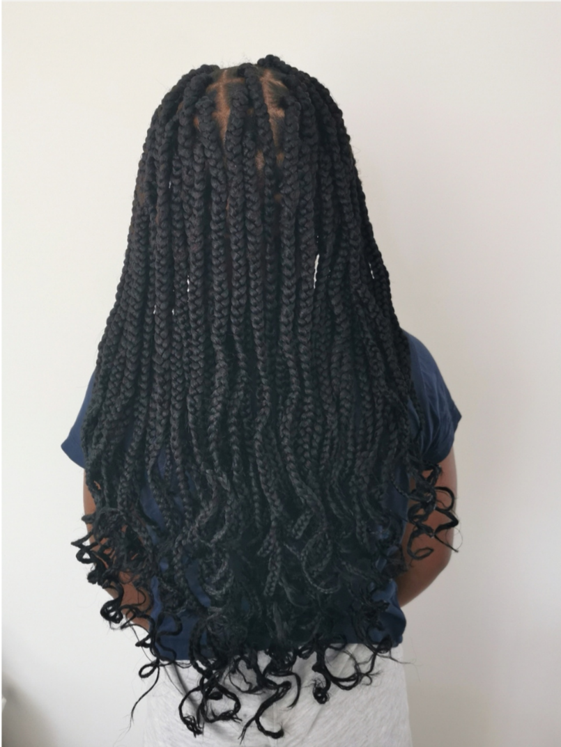 Back view of a person with long, thick, black twisted and curled hairstyle, wearing a navy shirt and light-colored pants, standing against a plain white wall.