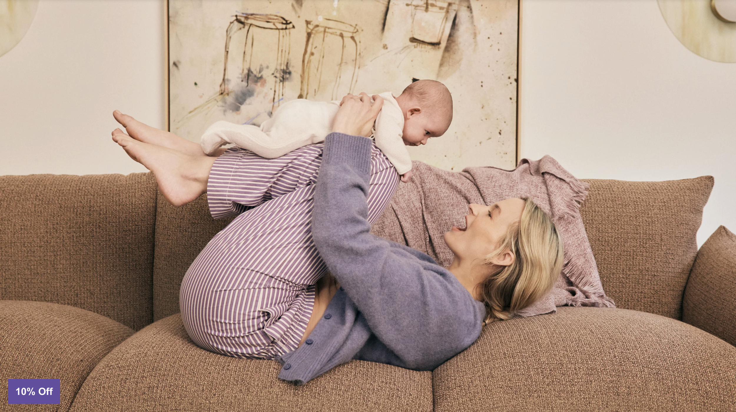 A woman and a child playing on a couch. The woman is lying down and holding a baby above her. The baby is dressed in light-colored clothes and is leaning forward towards the woman. The woman is wearing a purple cardigan and a striped skirt. The background features a wall with framed artwork.