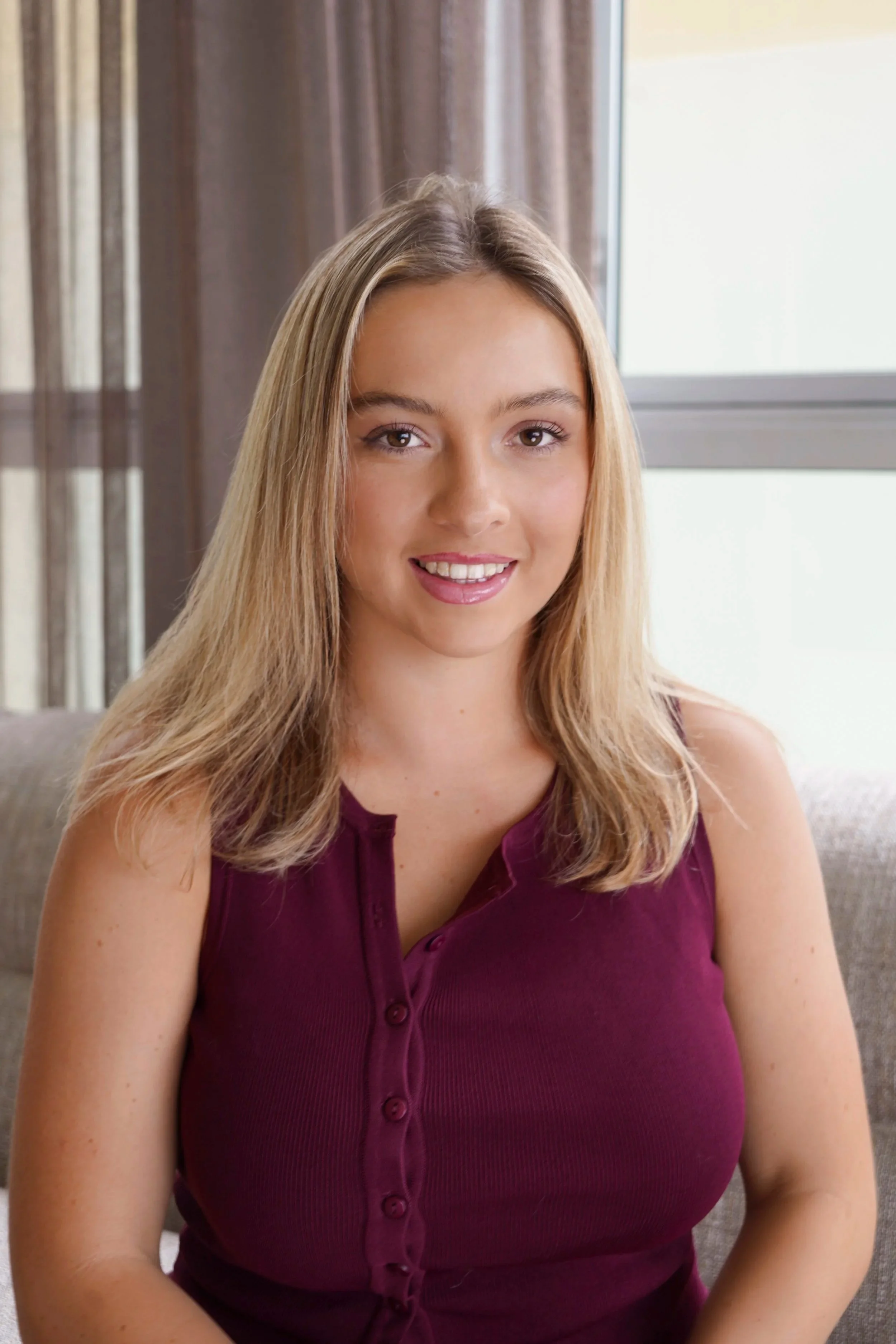A young woman with long, blonde hair, wearing a sleeveless purple top, smiling, sitting in front of a window with a brown curtain.