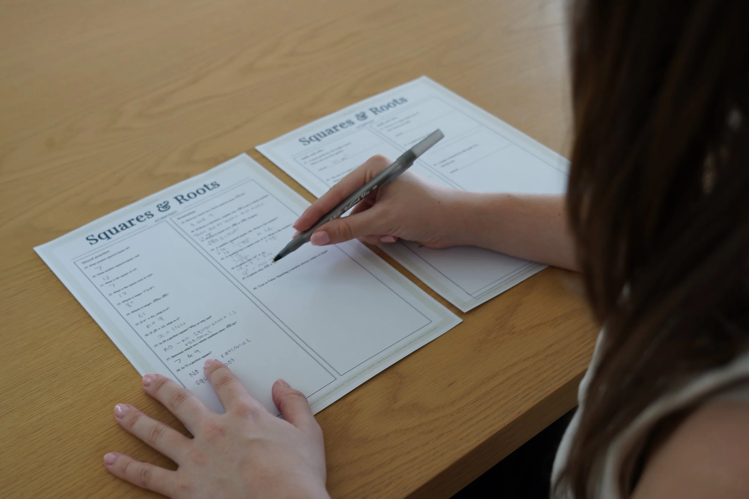 A person sitting at a wooden table, filling out a worksheet titled 'Squares and Roots' with a black pen.