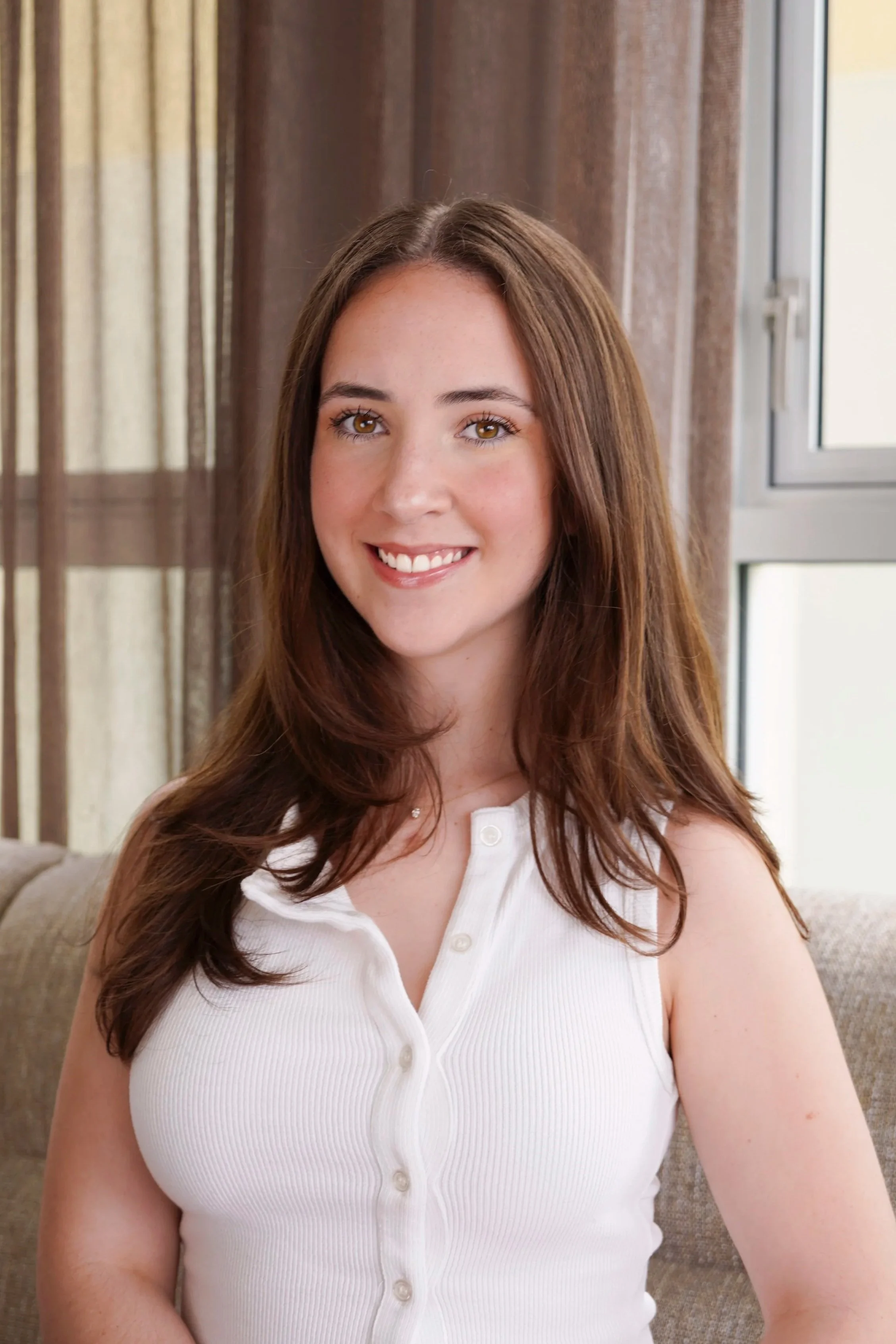 A young woman with long brown hair and hazel eyes smiling, wearing a sleeveless white top with buttons, sitting on a beige couch in front of a window with brown and beige curtains.