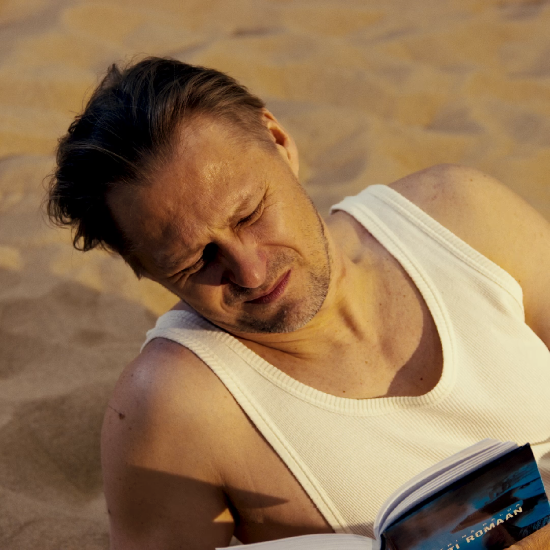 Close-up of a man lying on the ground, wearing a white tank top, with his eyes closed.