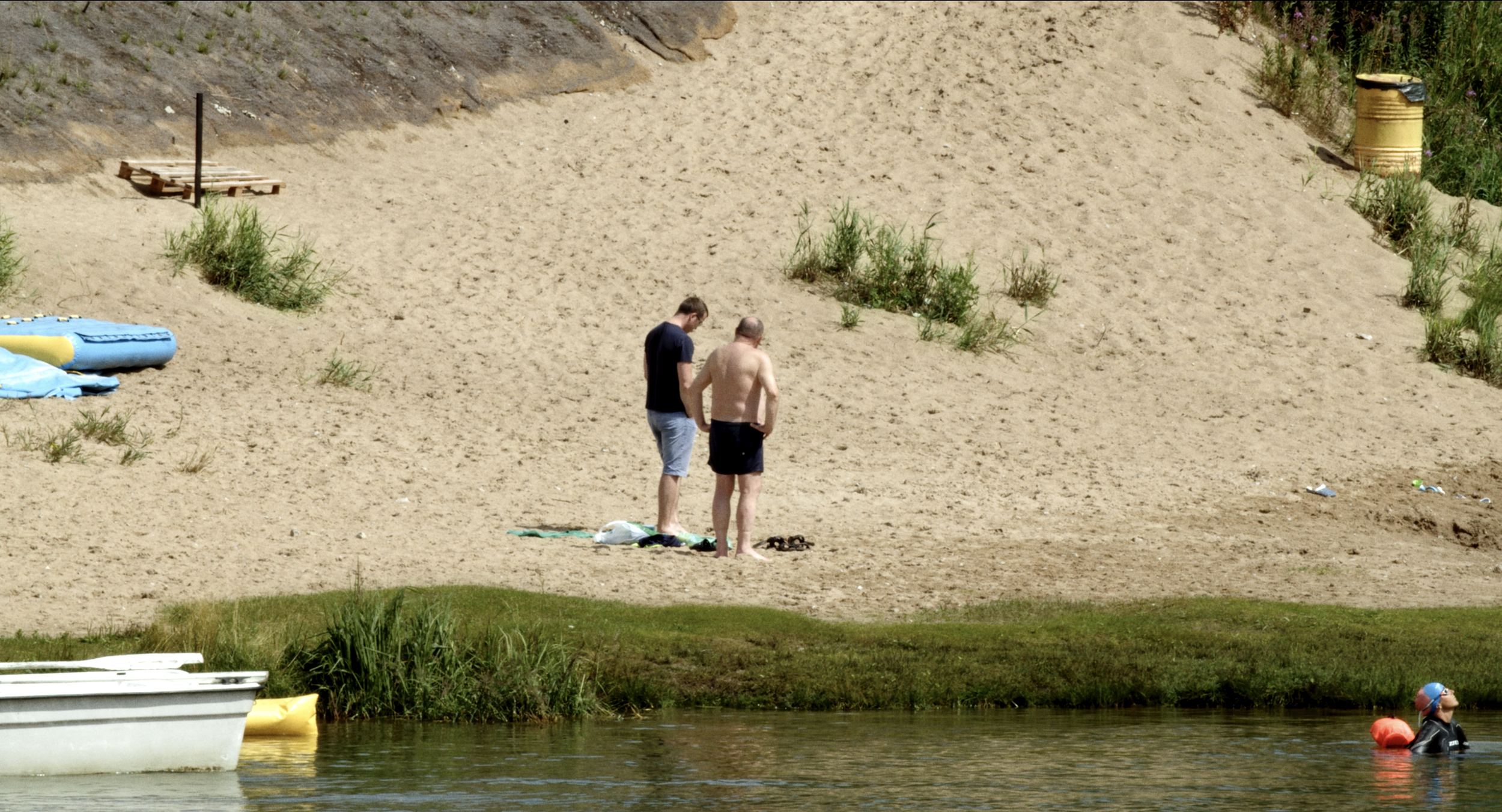 Two men standing on a sandy beach near a body of water, one shirtless and the other wearing a navy T-shirt, with floating toys and inflatable objects nearby, and vegetation along the shoreline.