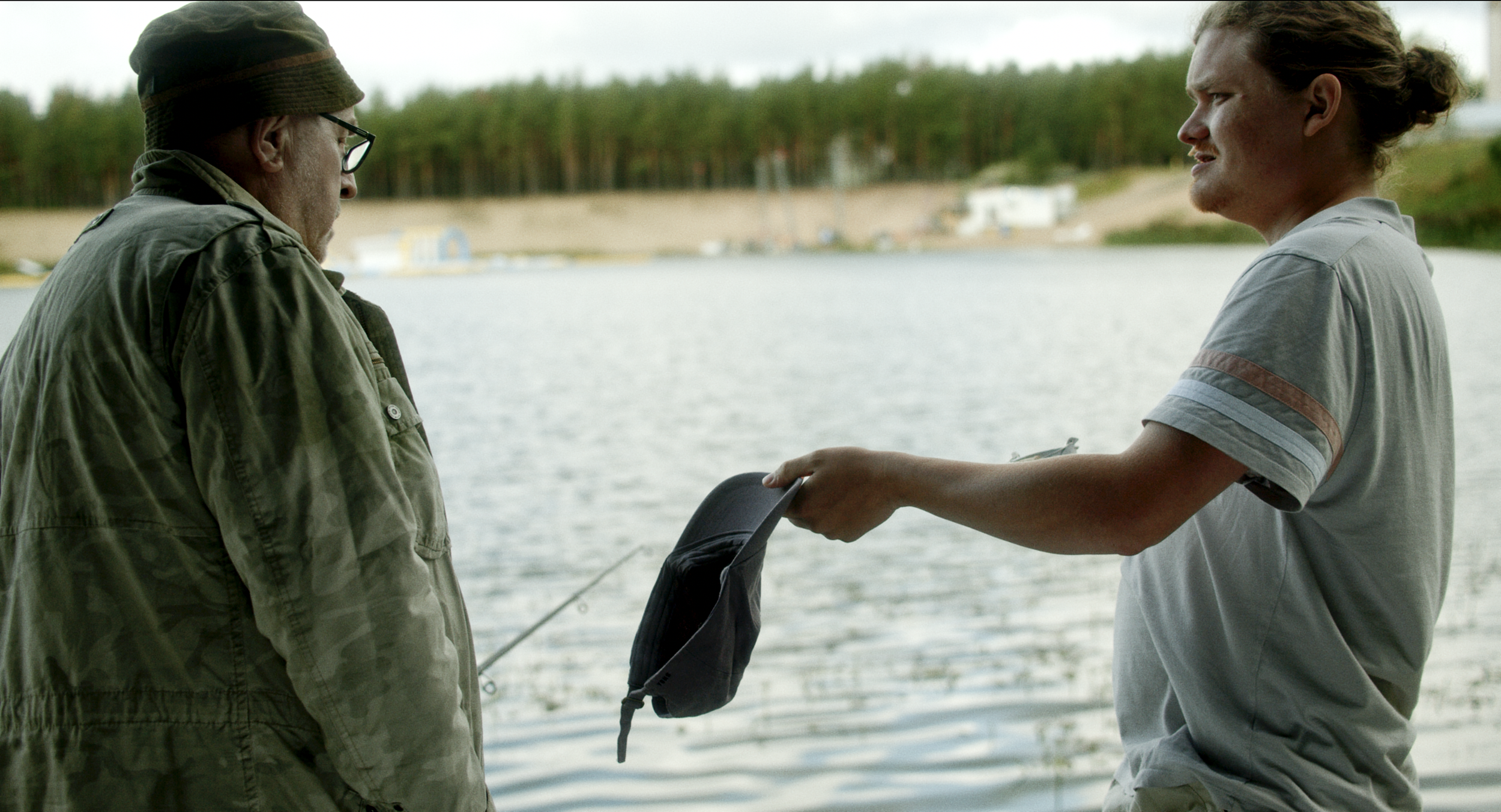 Two men standing near water, one in a green jacket and hat, the other in a gray t-shirt, one holding a backpack.