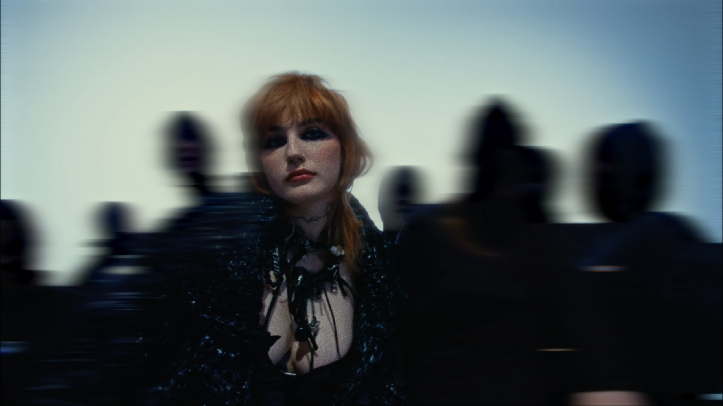 A woman with red hair and makeup, wearing a black sequined outfit with jewelry, is looking into the camera while reaching towards it, with a blurred background.