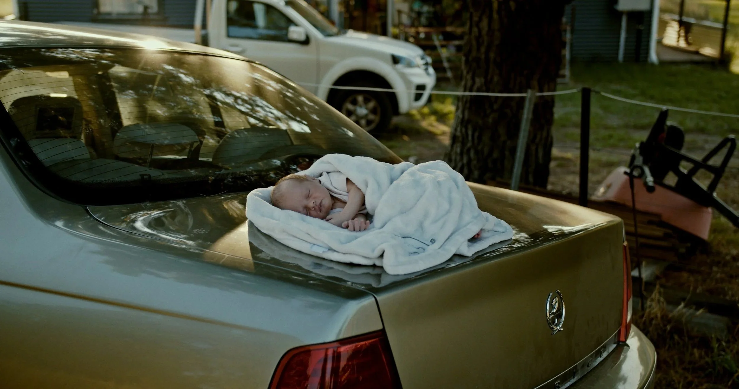 A newborn baby wrapped in a white blanket is sleeping on the hood of a yellow car outside, with a white truck in the background and trees surrounding the area.