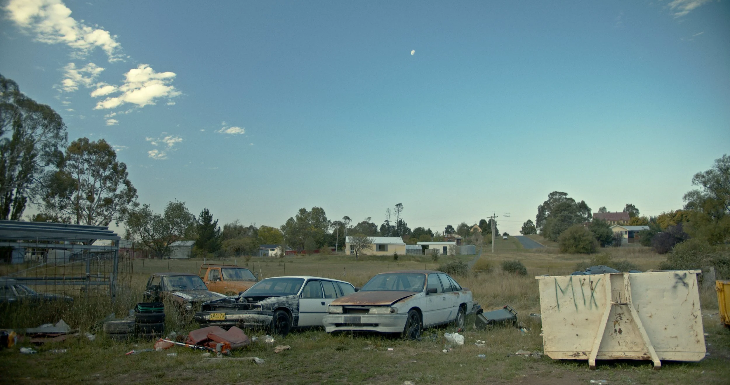 A rural area with several parked and abandoned cars, some rusted, with trees and houses in the background under a blue sky with some clouds and the moon visible.