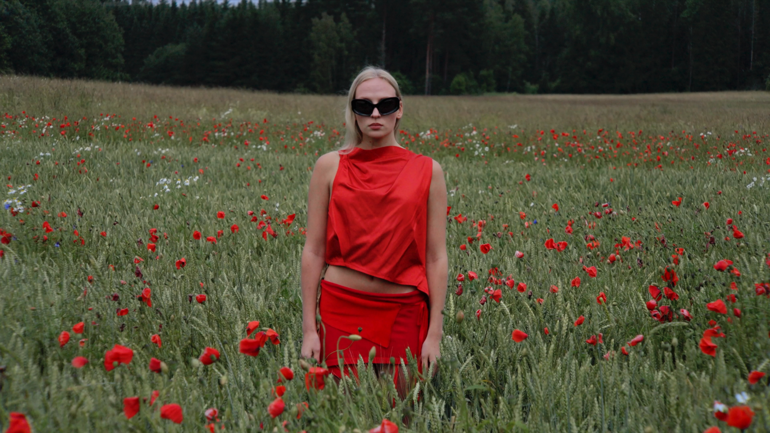 A woman with blonde hair wearing oversized black sunglasses and a red stylish outfit walking through a field of red poppies with a forest in the background.