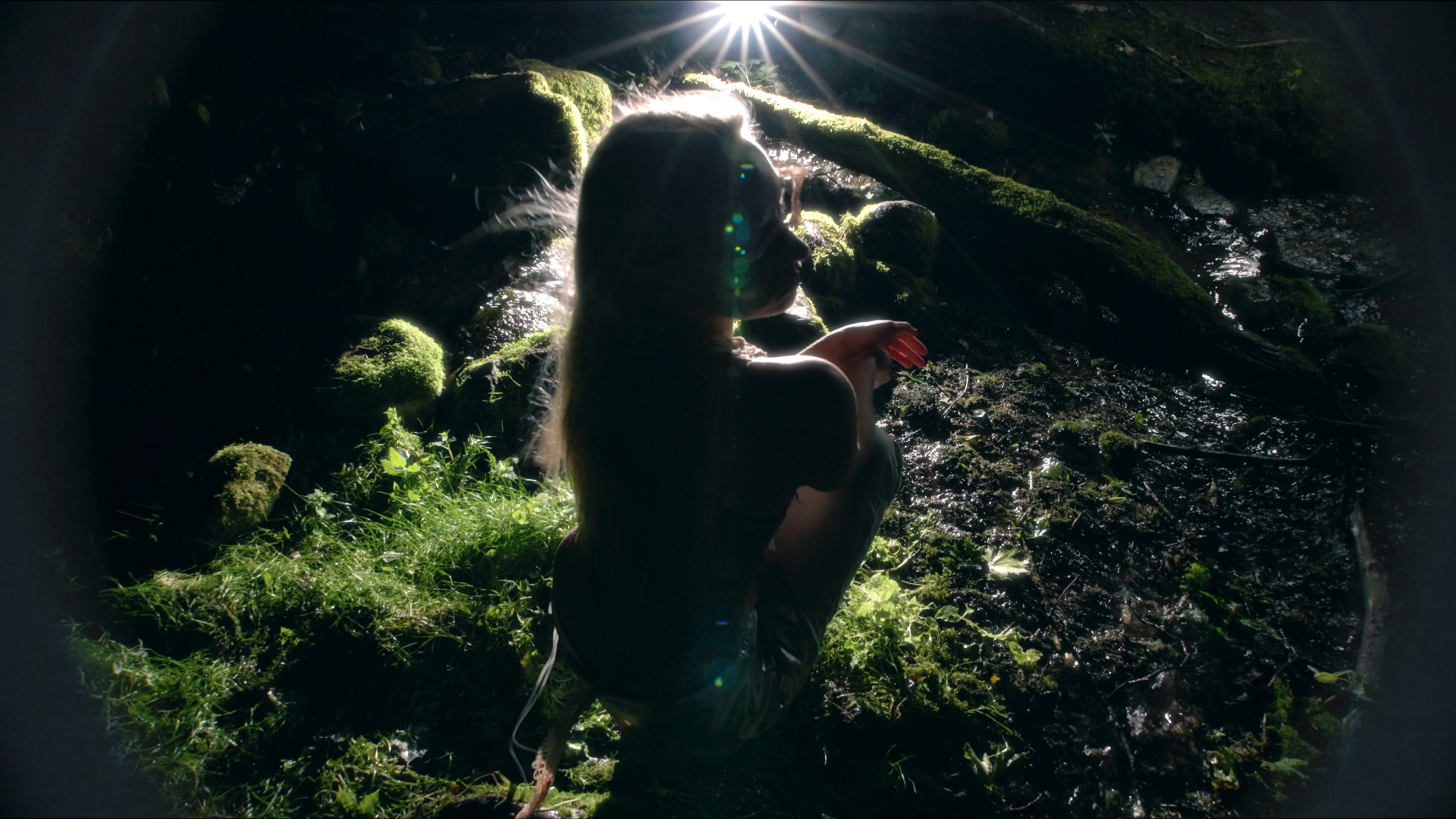 A woman with long blonde hair, sitting in a lush mossy forest near a small stream, backlit by sunlight filtering through the trees.