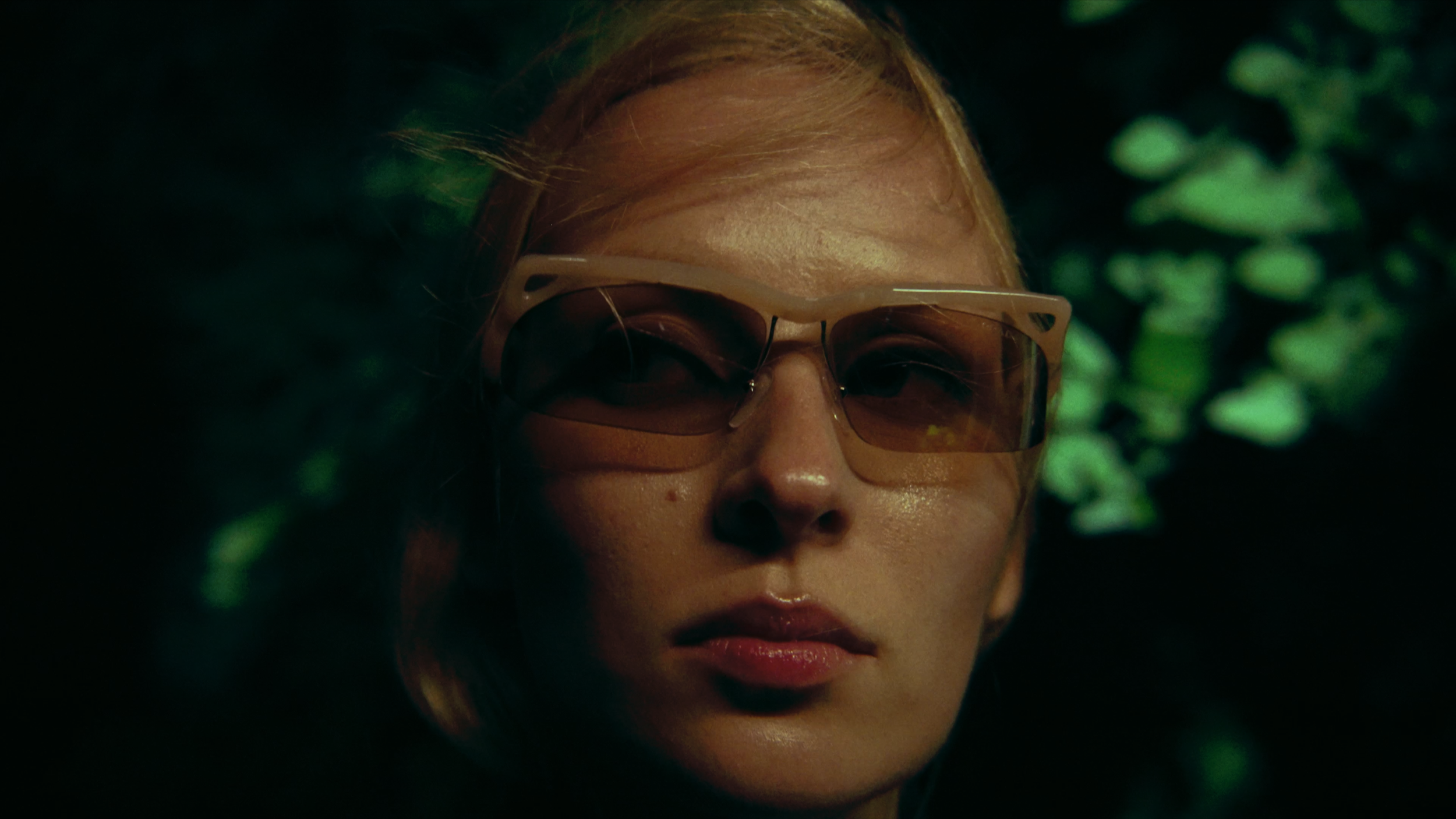 Close-up of a woman's face wearing large beige sunglasses, with blonde hair, surrounded by dark green foliage.