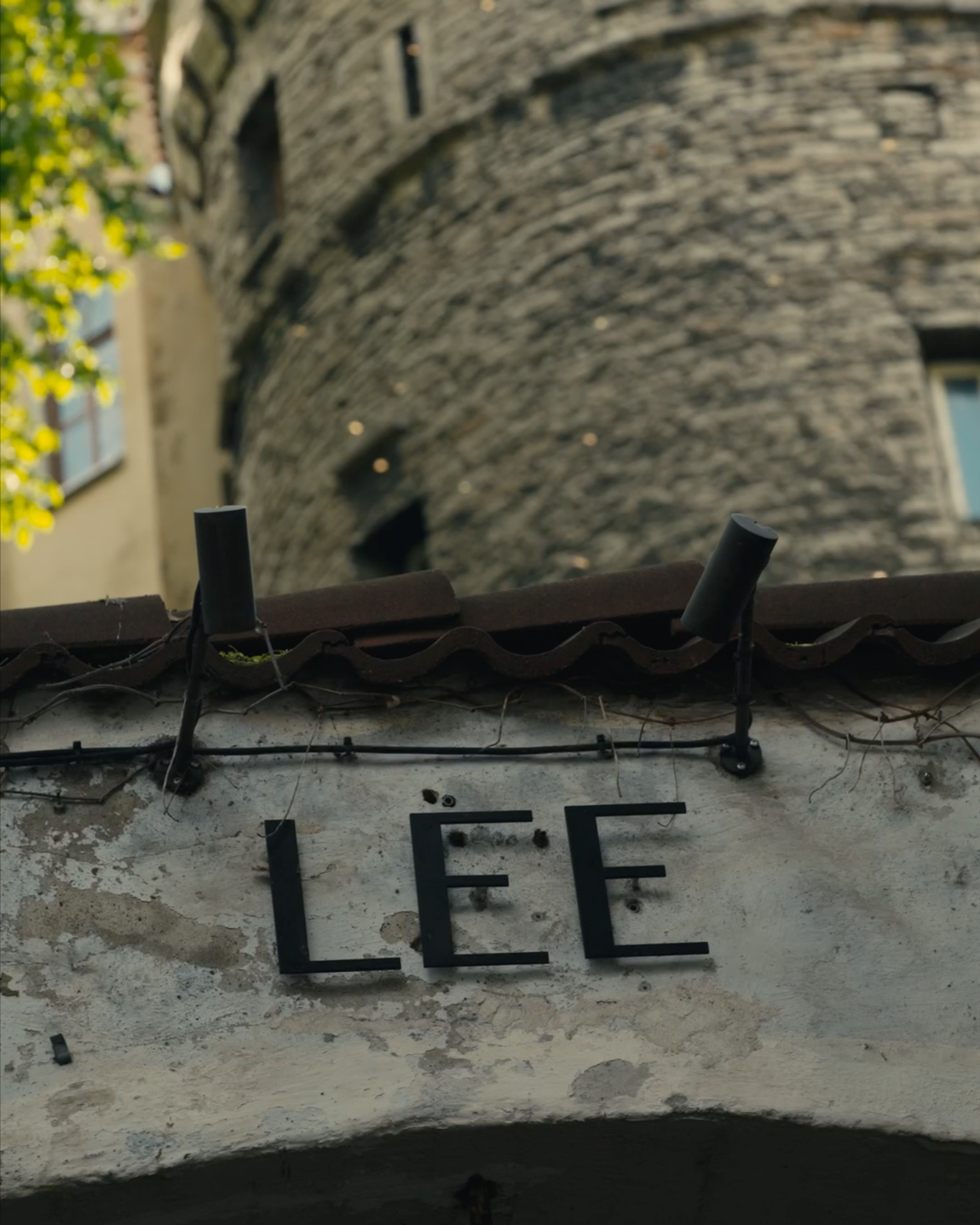 Close-up of a weathered sign with the letters 'LEE' mounted on a textured wall with black wires and small black lights above, with a blurred stone building and tree in the background.