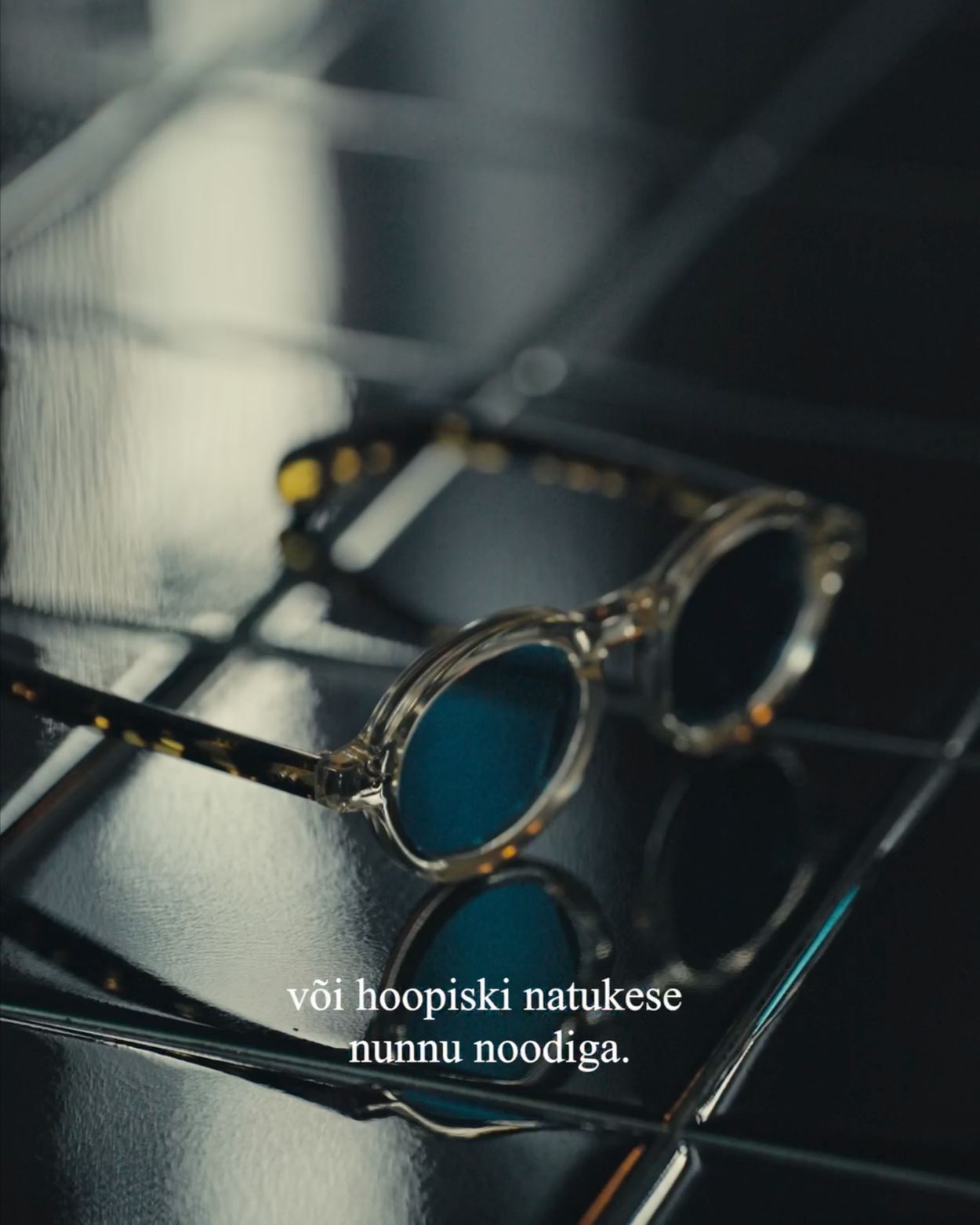 Close-up of blue-tinted round sunglasses on a metal rack with a metallic surface in the background. Overlaid text reads, "või hoopiski natukese nunnu noodiga."