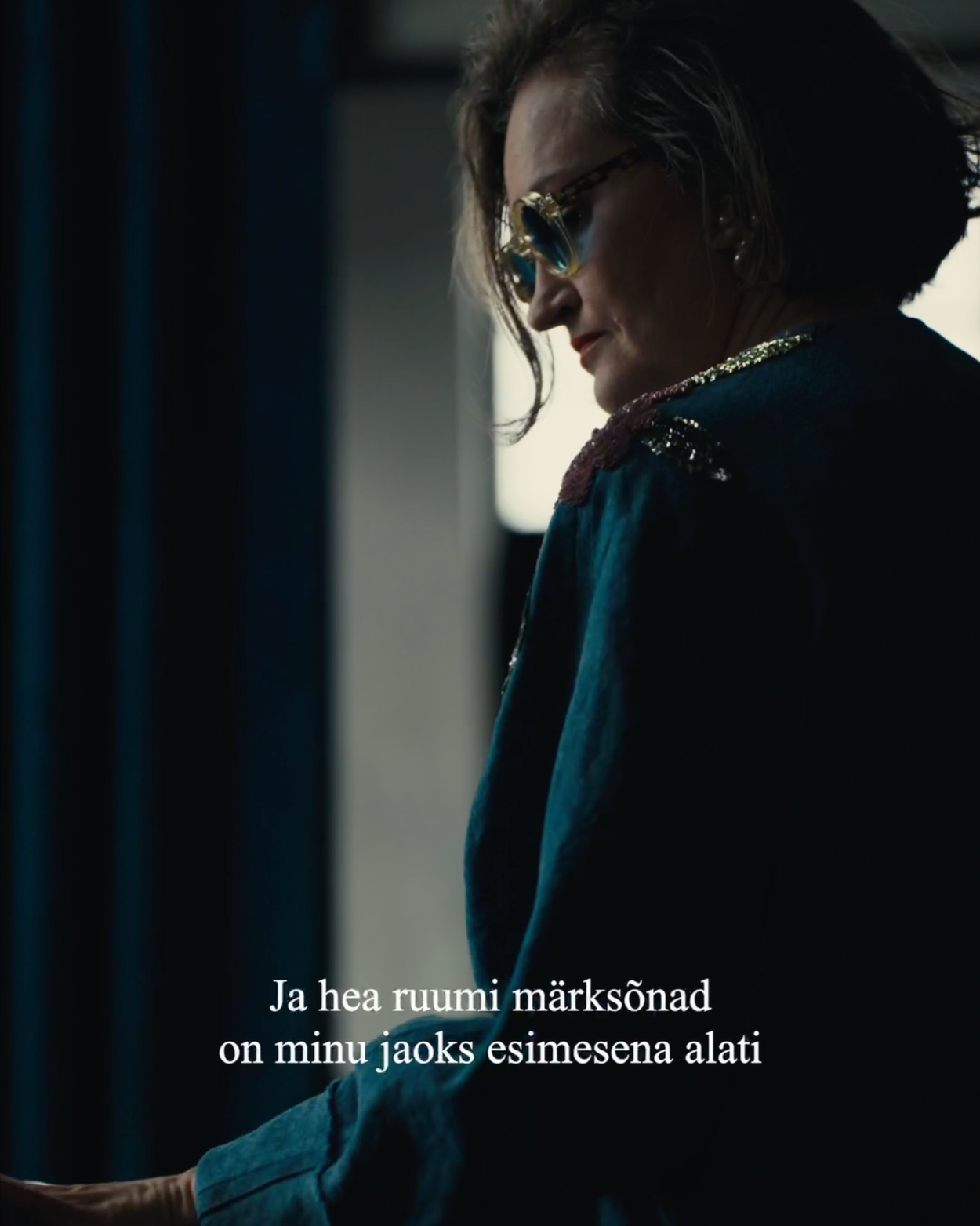 A woman with short, styled hair wearing sunglasses and a dark jacket, standing indoors near a window with a quote in Finnish at the bottom.