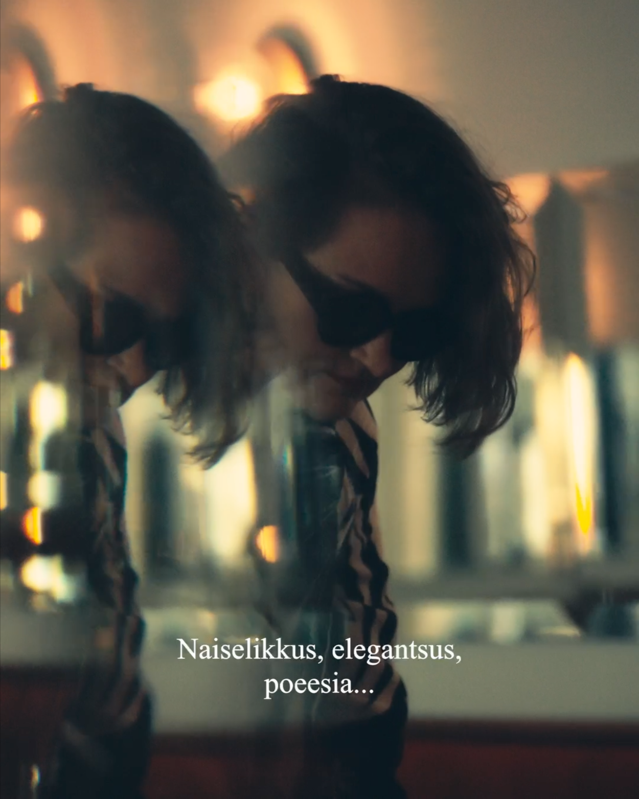 A woman with dark, wavy hair and wearing sunglasses leaning against a metallic surface with warm, blurry lights in the background.
