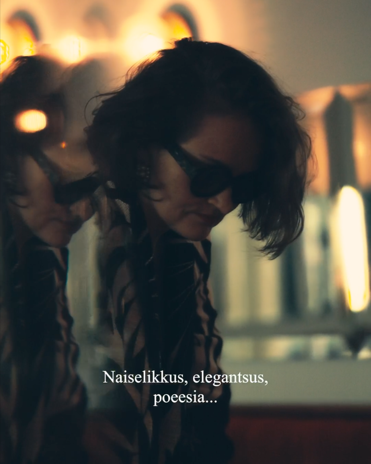 Two women with dark, curly hair and sunglasses leaning in close together against a blurred background with warm lighting. Subtitles read: "Naiselikkus, elegantsus, poeasiao..."