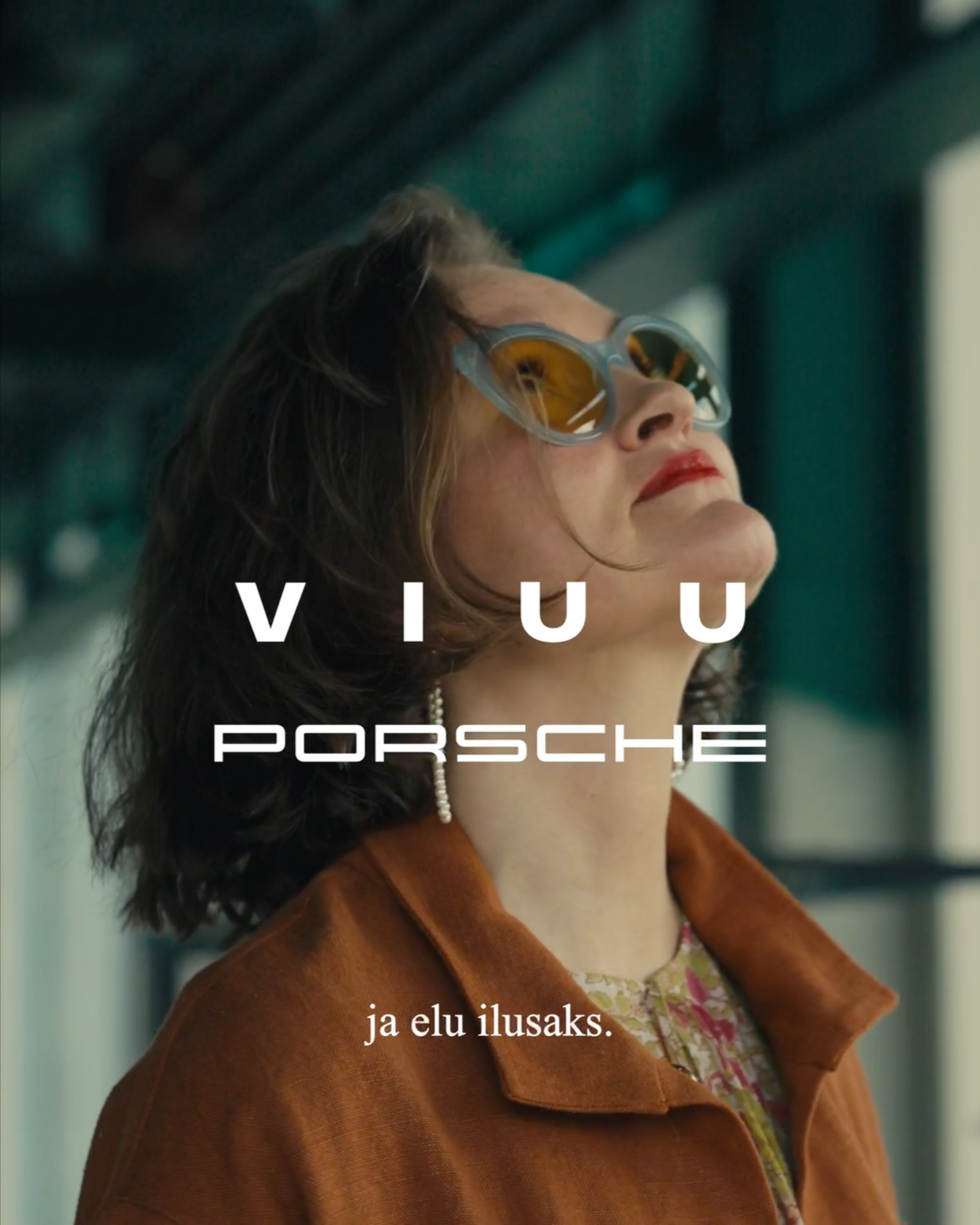 A woman with dark hair, wearing yellow sunglasses, a pearl necklace, and a brown coat, is looking up and smiling inside a bus or train.