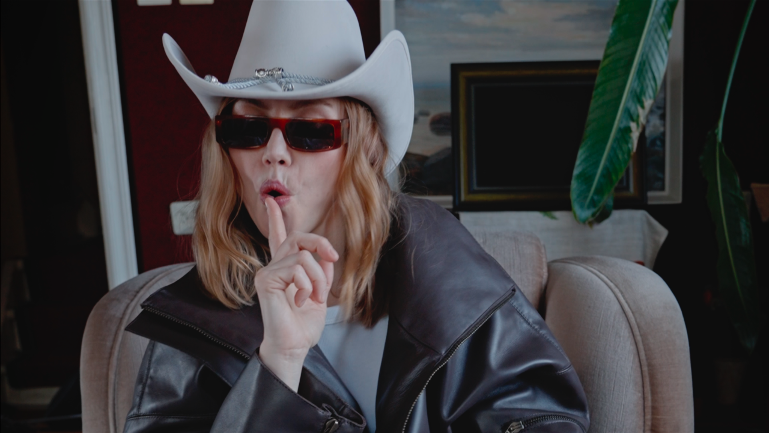 Woman wearing a white cowboy hat, red sunglasses, and a leather jacket making a shushing gesture with her finger.