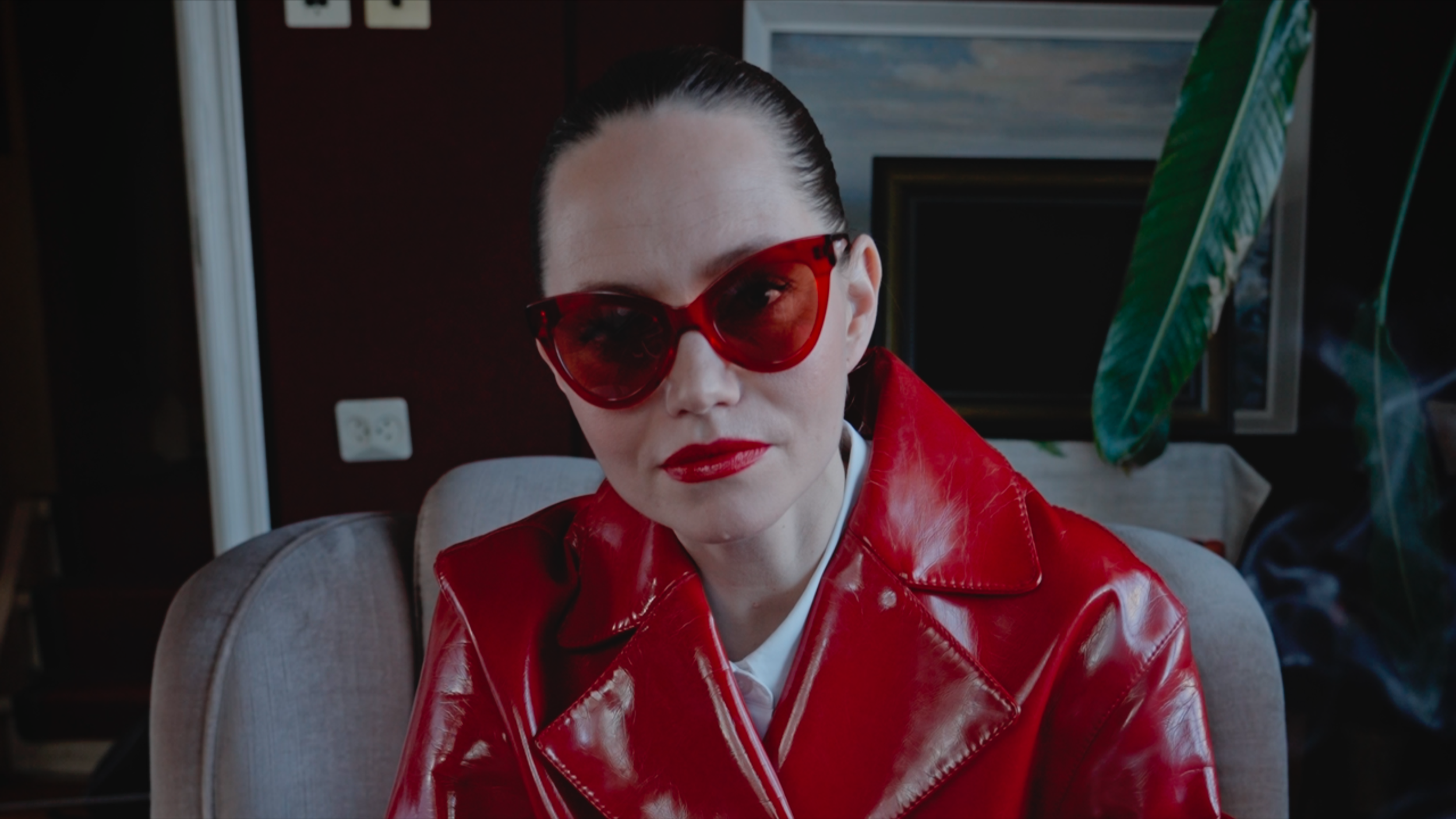 A woman wearing red oversized sunglasses, red lipstick, and a shiny red leather jacket, sitting indoors near a large green plant.