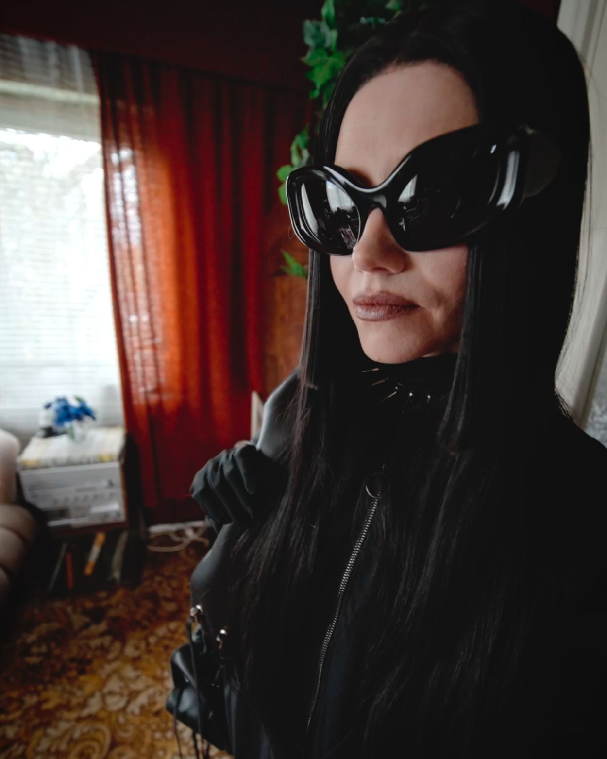 Woman wearing large black sunglasses and a black leather jacket, standing indoors near a window with red curtains and a plant in the background.