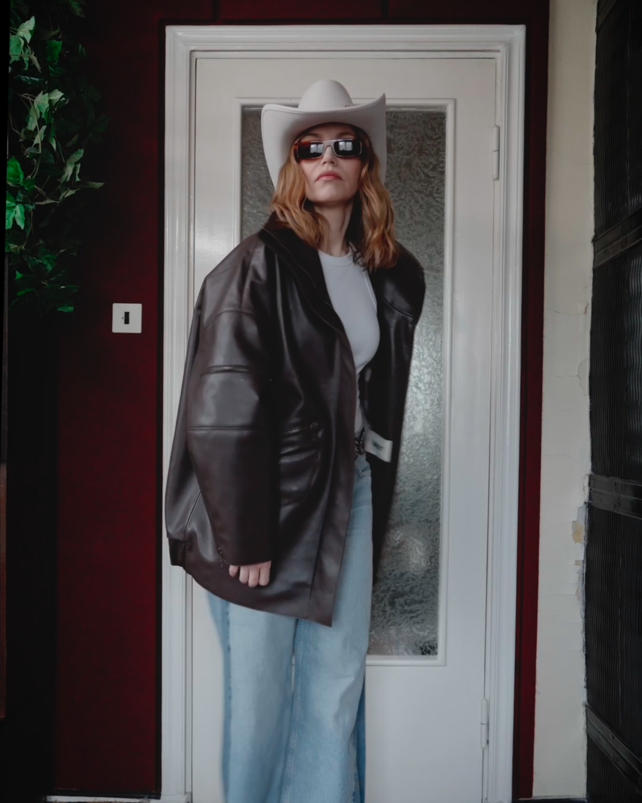 Woman wearing a white cowboy hat, dark sunglasses, a black leather jacket, a white shirt, and loose light blue jeans standing in front of a door with frosted glass.