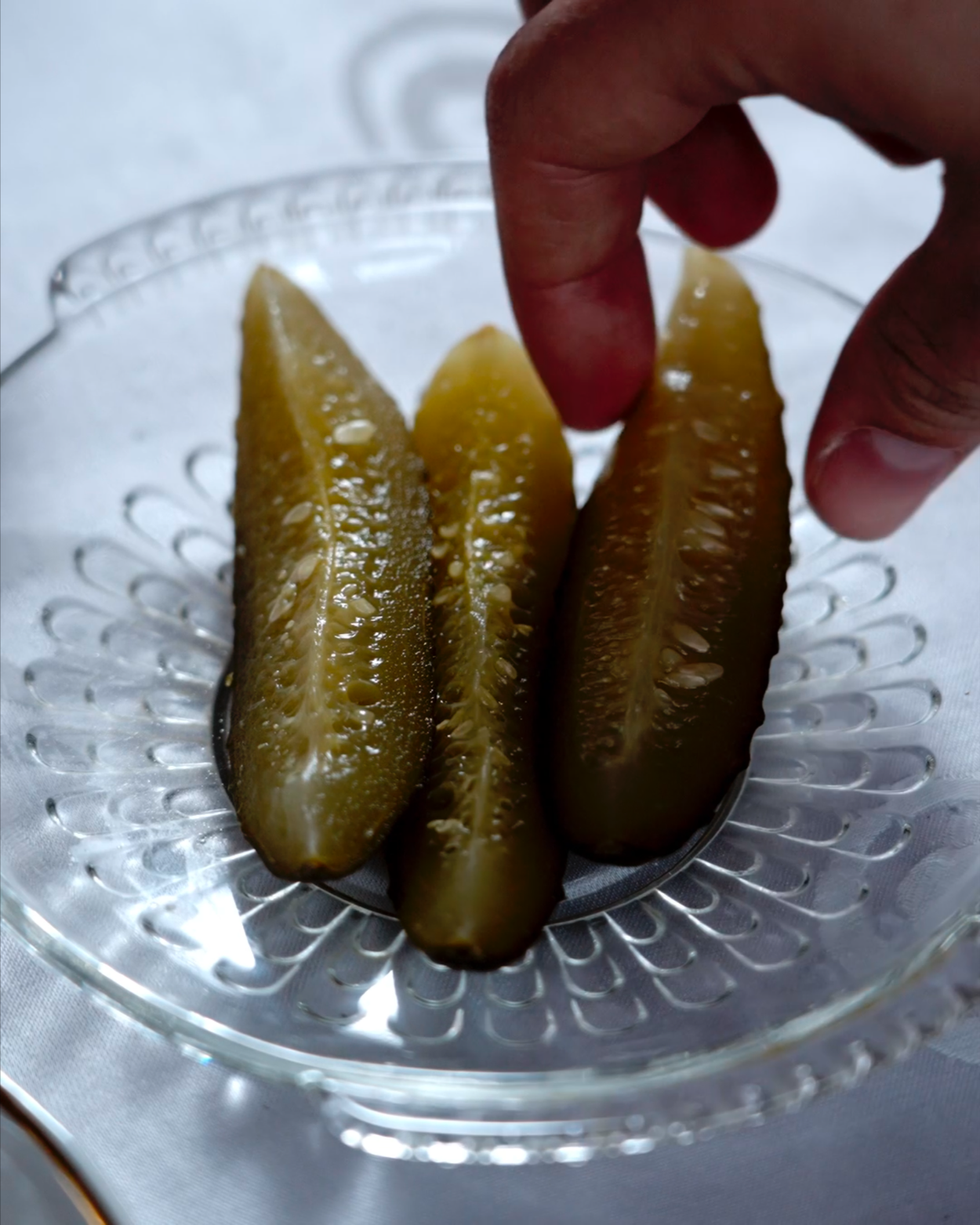 Three slices of pickles on a glass plate with a decorative pattern, with a person's hand holding the top of one pickle.