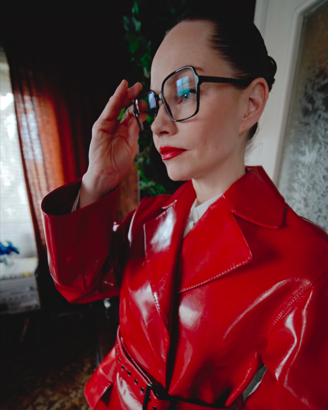 Woman with dark hair, red lipstick, glasses, wearing a shiny red vinyl trench coat, adjusting her glasses indoors.