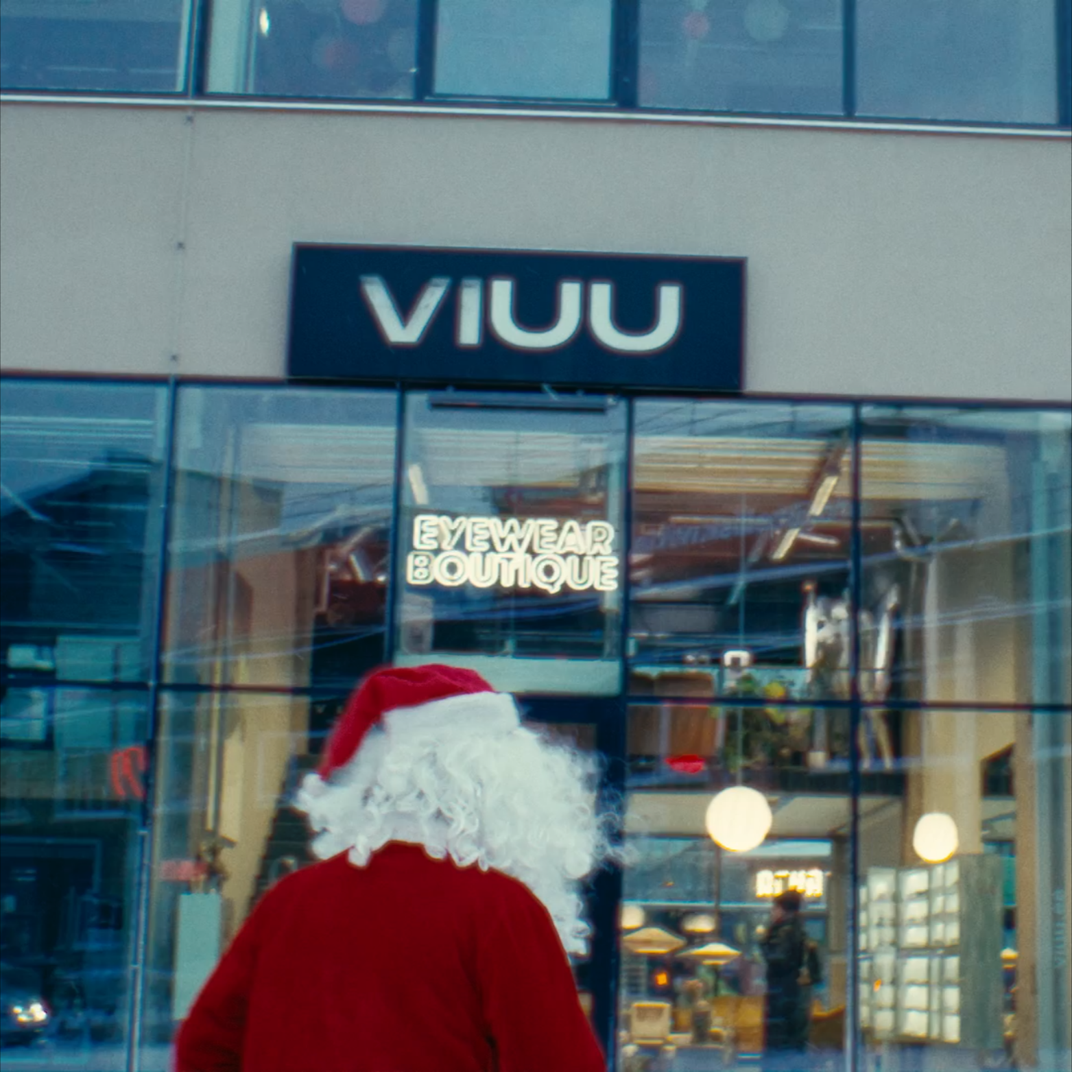The image shows the back of a person dressed as Santa Claus, facing store windows with signs that say "VIUU" and "EYEWEAR BOUTIQUE." There are reflections of the store's interior, lights, and other people outside.