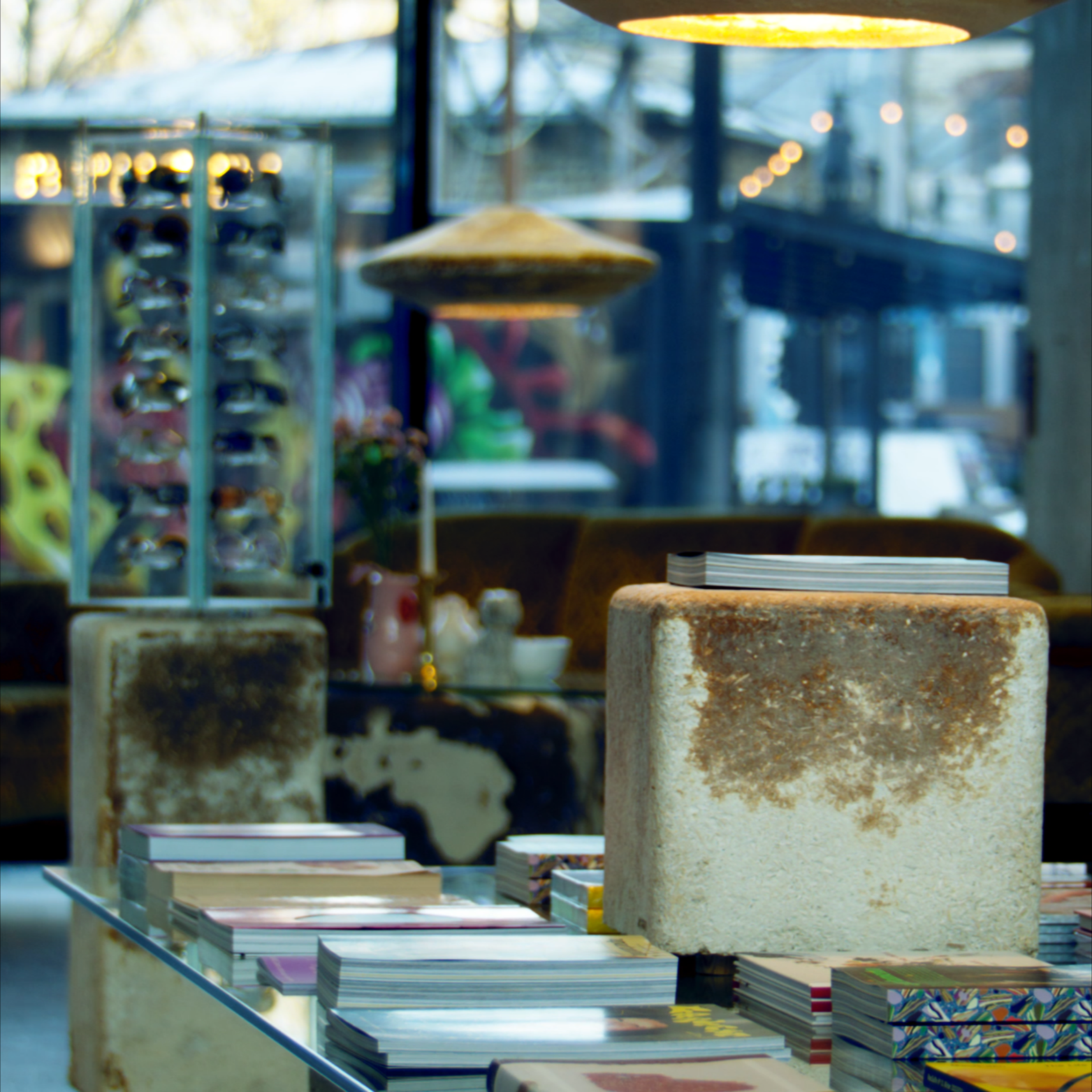 Display of various books and magazines on a glass table with stone blocks in an indoor space, with blurred windows and string lights in the background.